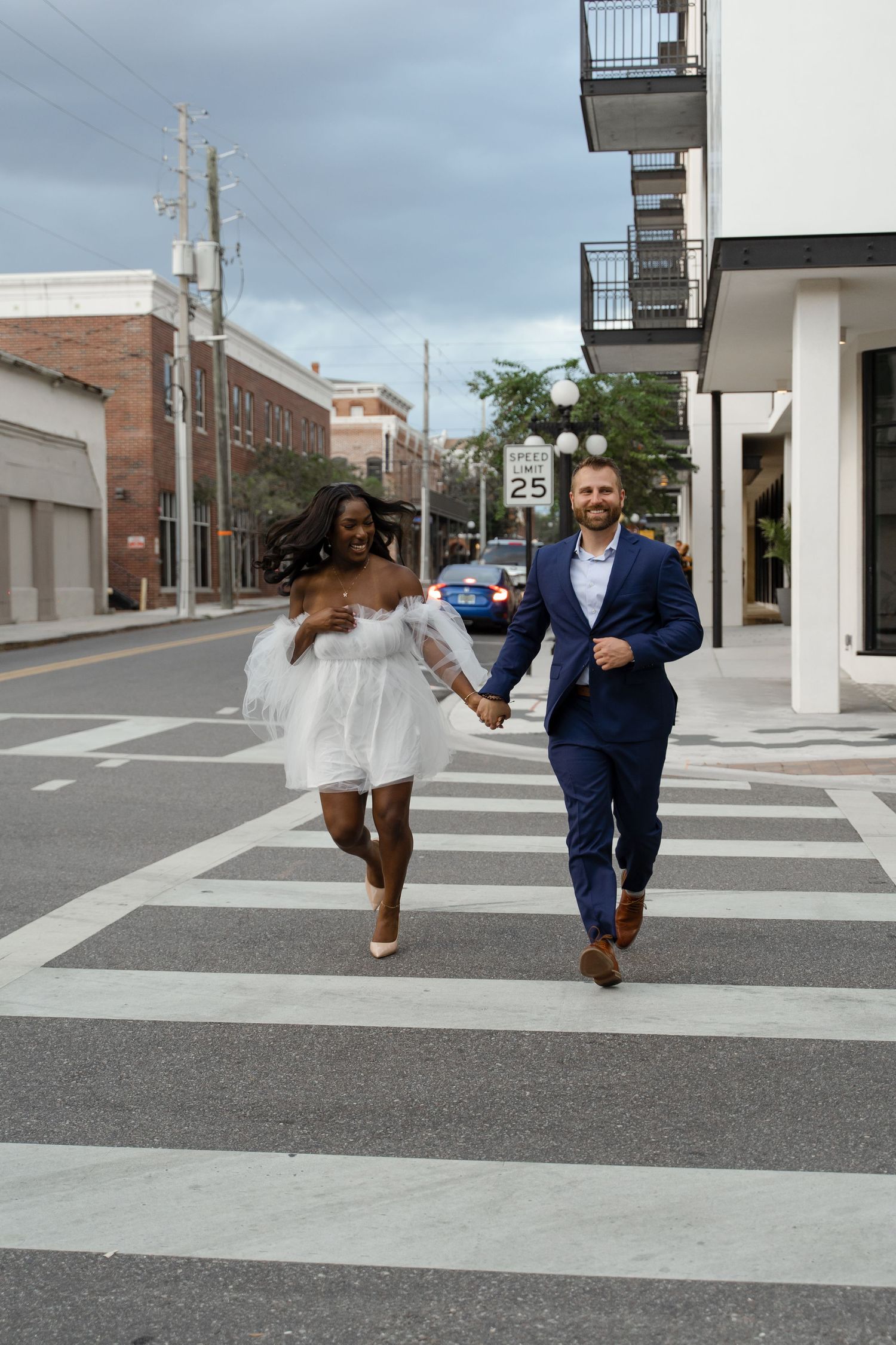 Joyful interracial couple in wedding attire running hand-in-hand across a city crosswalk.