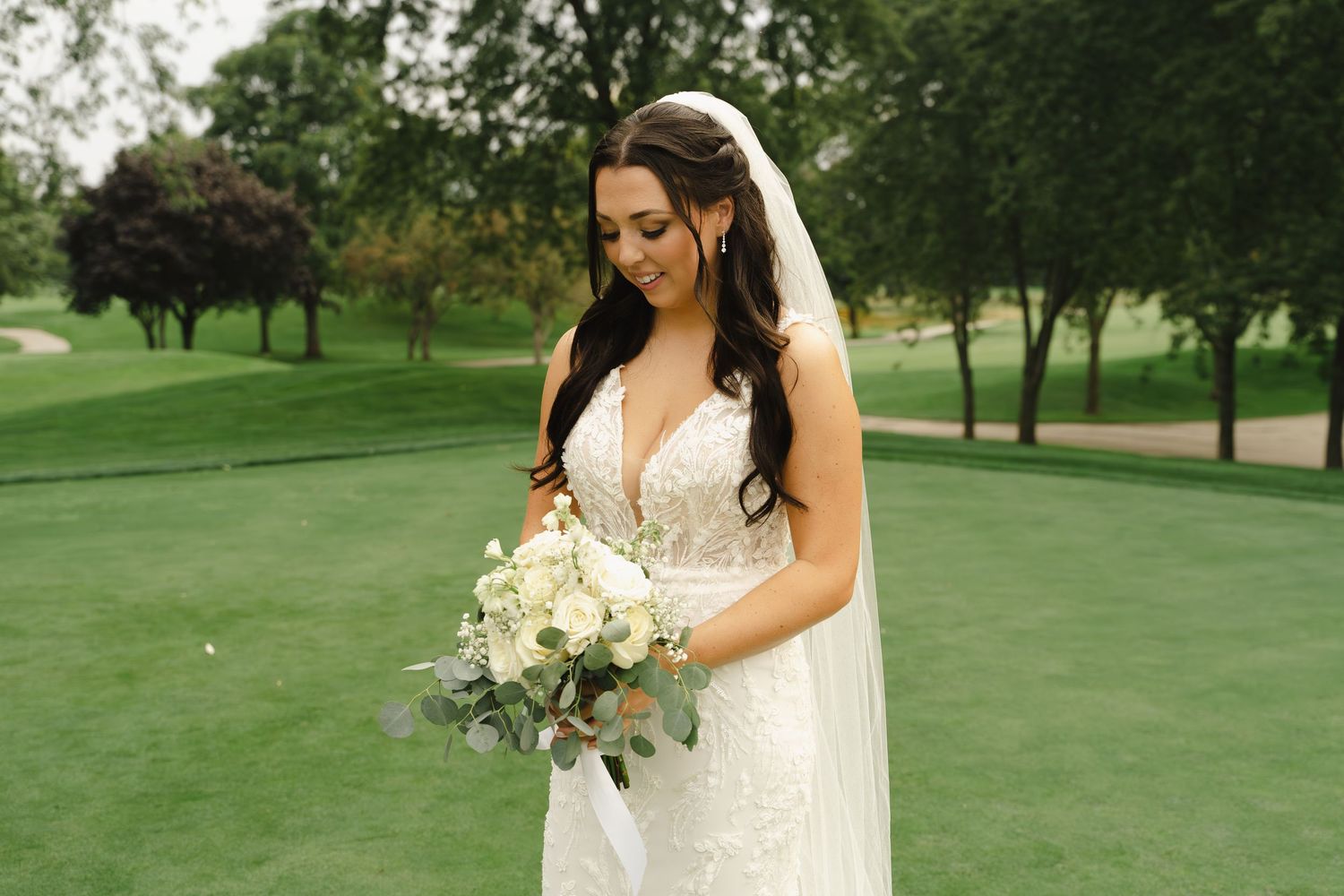 Bride in lace gown holding white bouquet while standing on a golf course green.