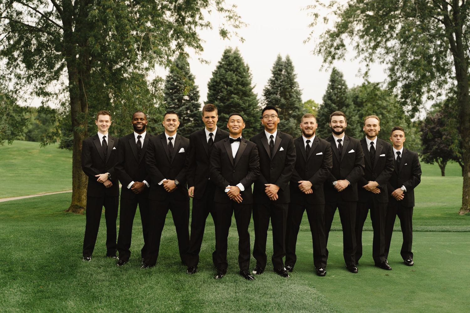 Groom poses with groomsmen all dressed in matching black tuxedos on golf course.