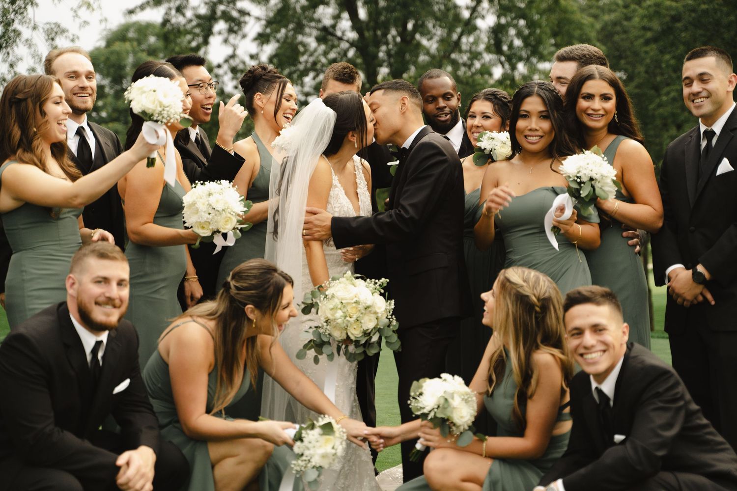 Smiling wedding party in sage green dresses and black suits gathered outdoors with bouquets.