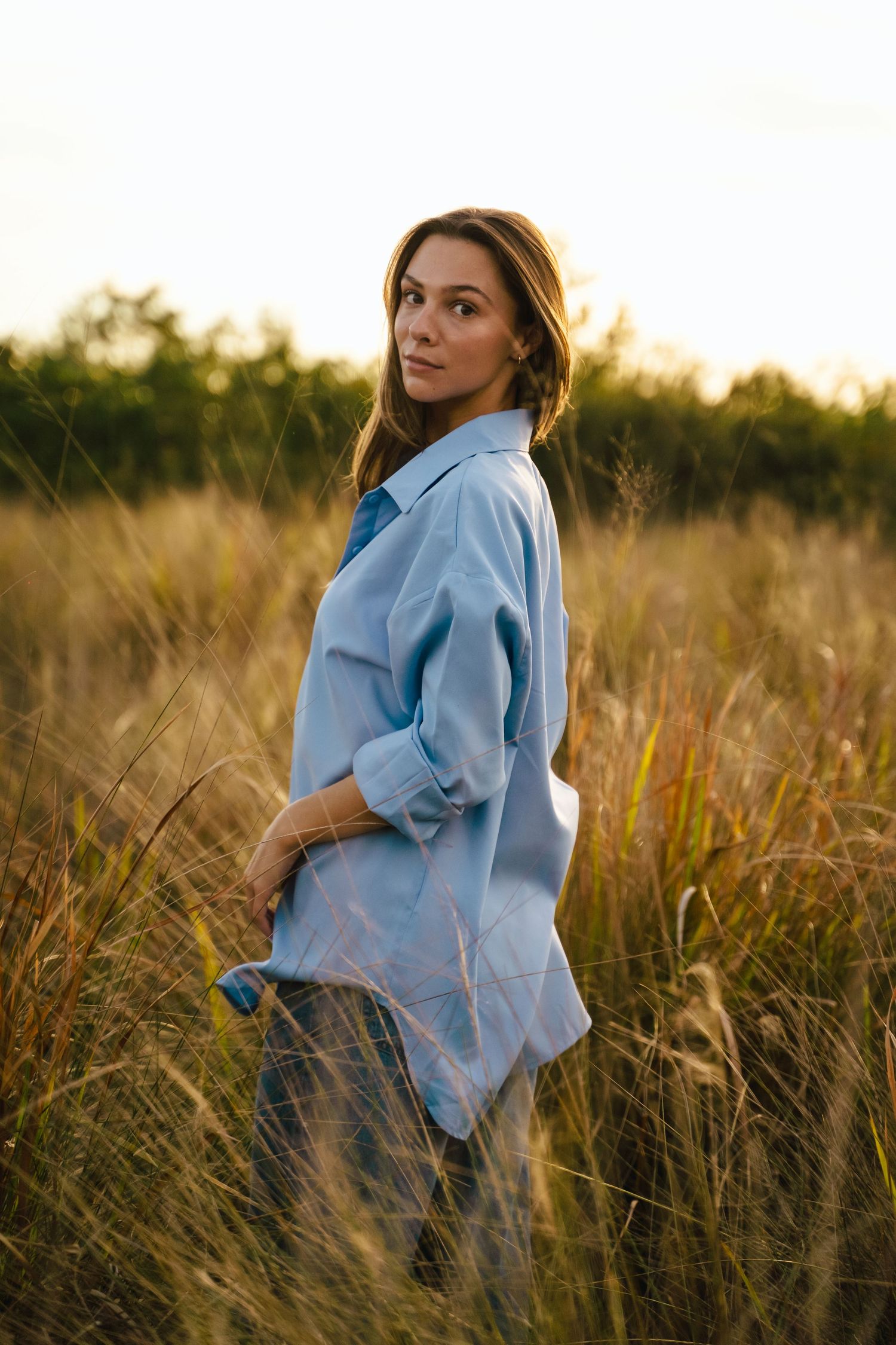 Woman in oversized blue shirt standing in golden tall grass field during warm sunset light.