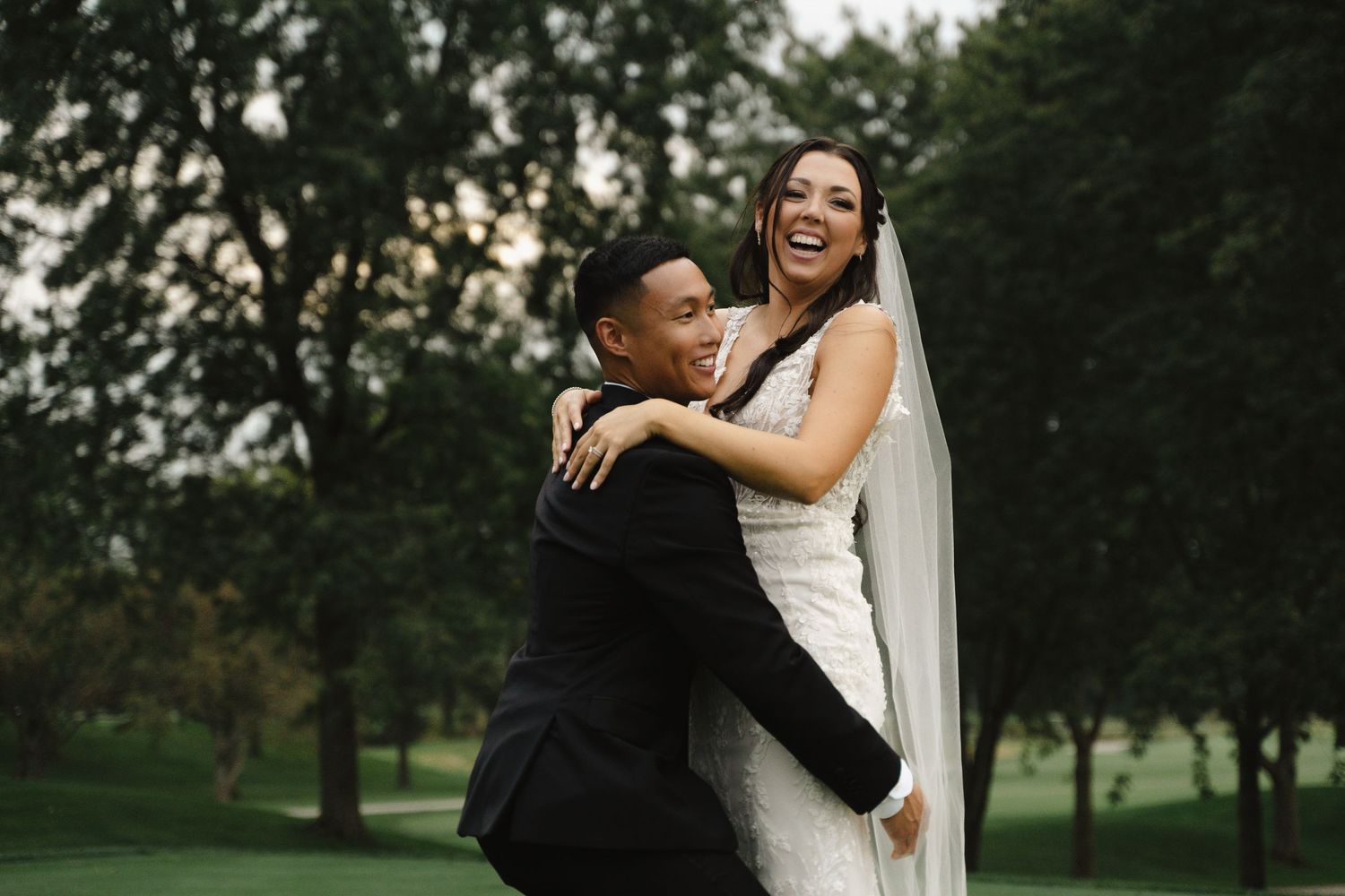 Joyful groom lifting laughing bride in white wedding gown and veil outdoors among green trees.