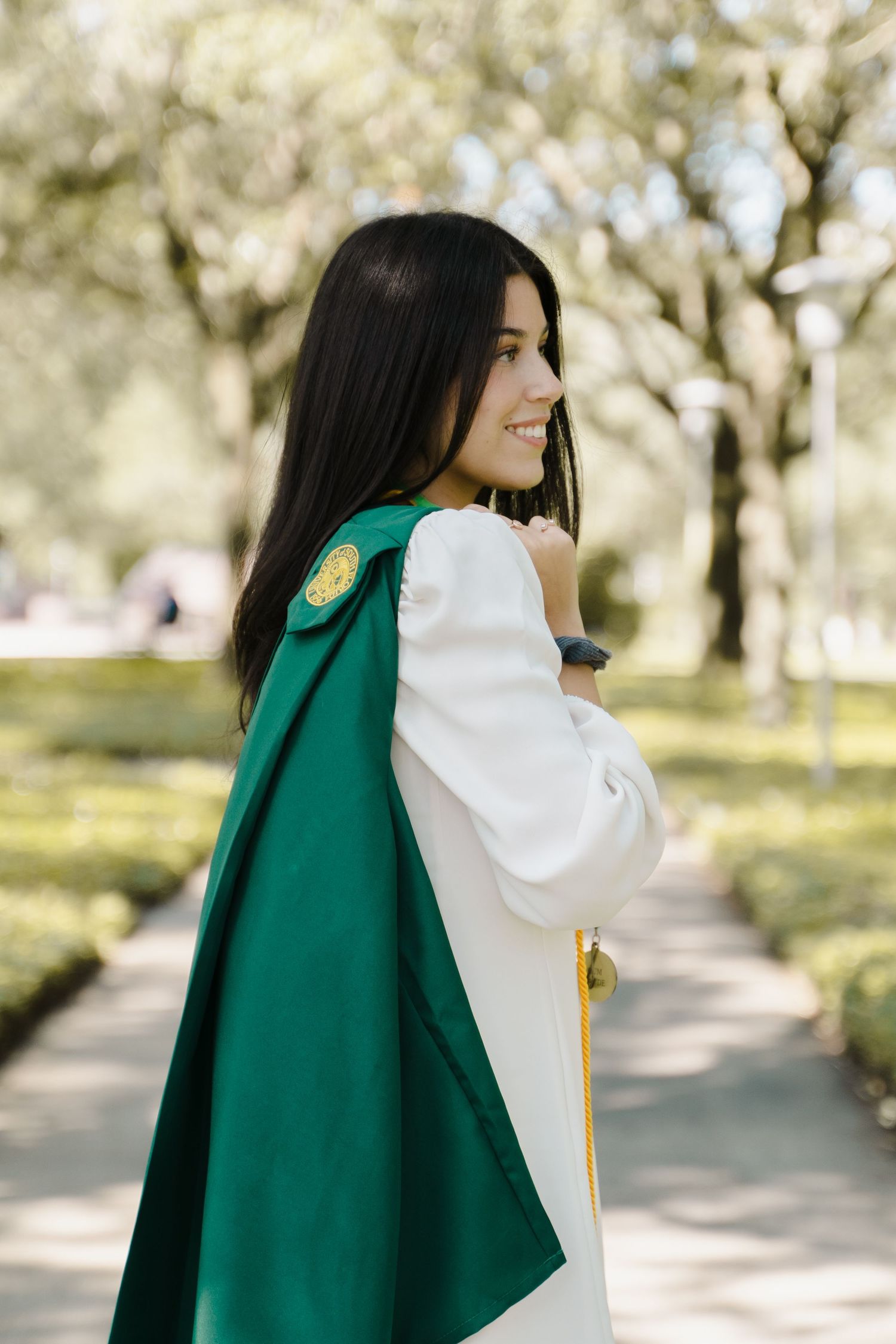 Smiling young woman in green graduation regalia poses on sunny campus pathway with bokeh background.