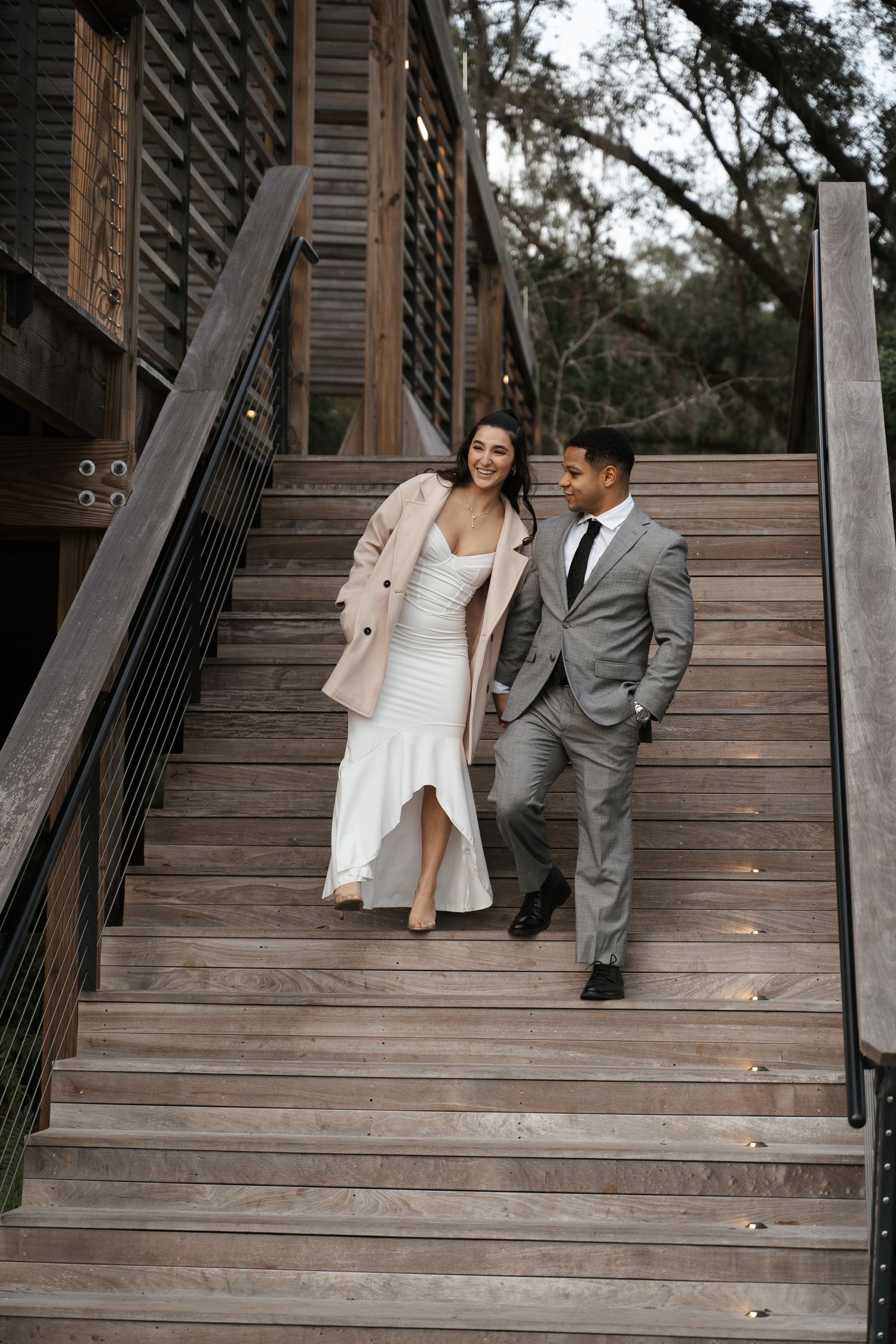 Smiling couple descending wooden stairs, woman in white dress with beige coat and man in gray suit.