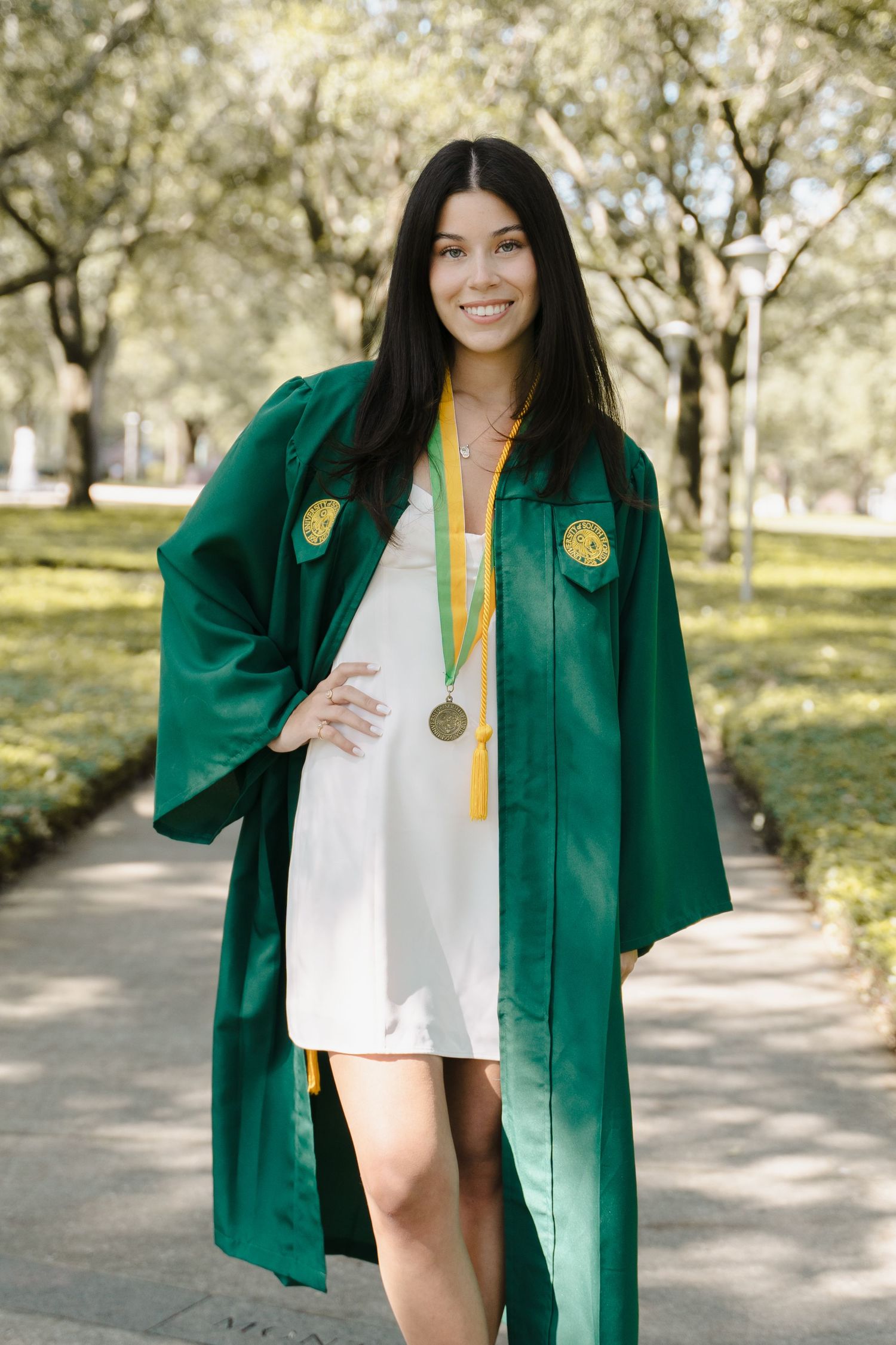Smiling woman in green graduation gown and medal poses outdoors on a tree-lined campus walkway.