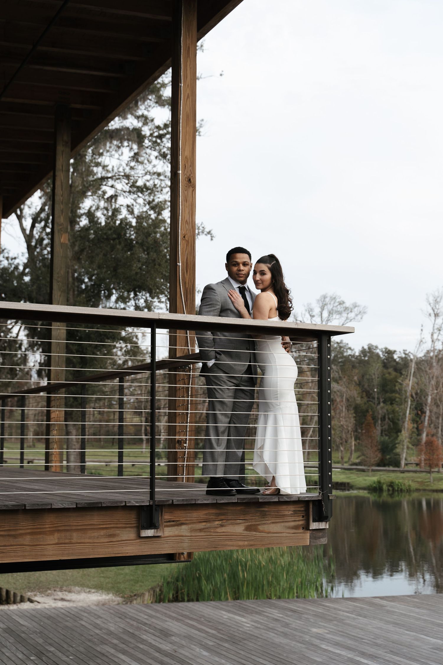Elegant wedding couple poses on a wooden deck balcony with a peaceful pond and lush greenery in the background.