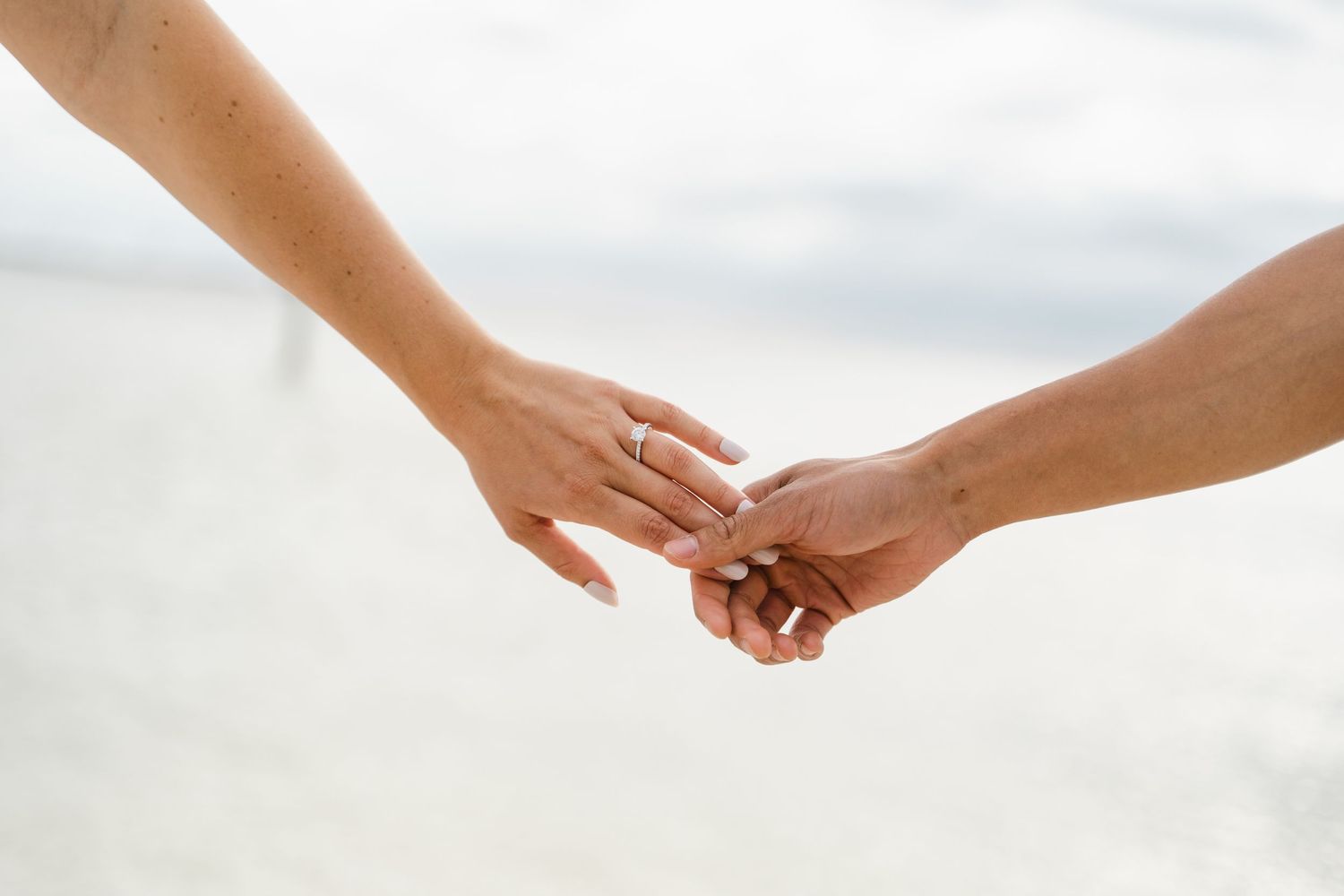 Couple holding hands on a beach with wedding rings visible, romantic engagement or wedding photo by the ocean.