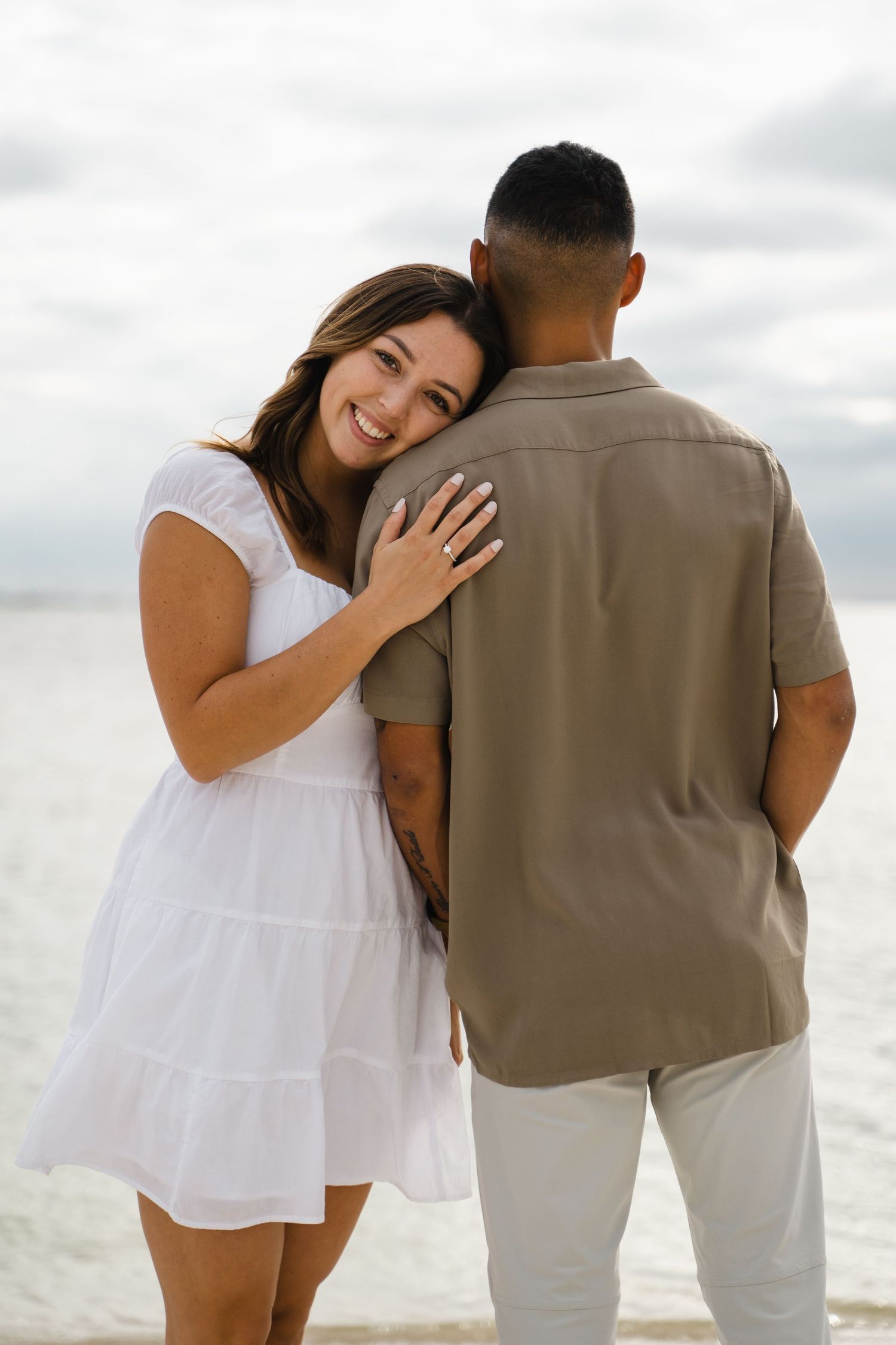A happy interracial couple hugs on a beach, woman in white sundress leaning on mans shoulder smiling.