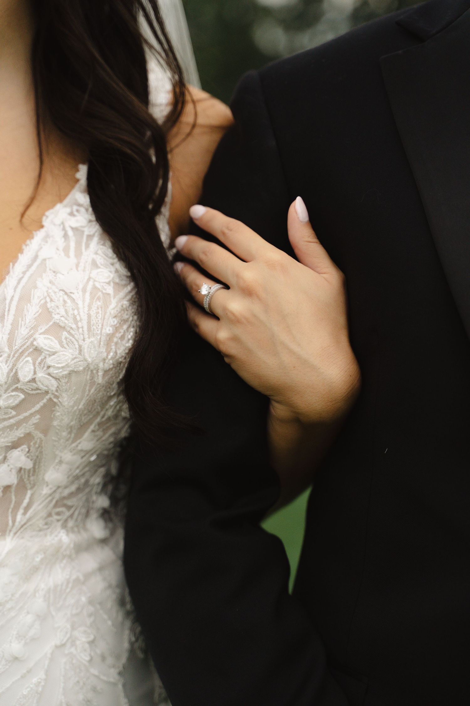 Close-up of bride's hand with diamond engagement ring resting on groom's black suit.