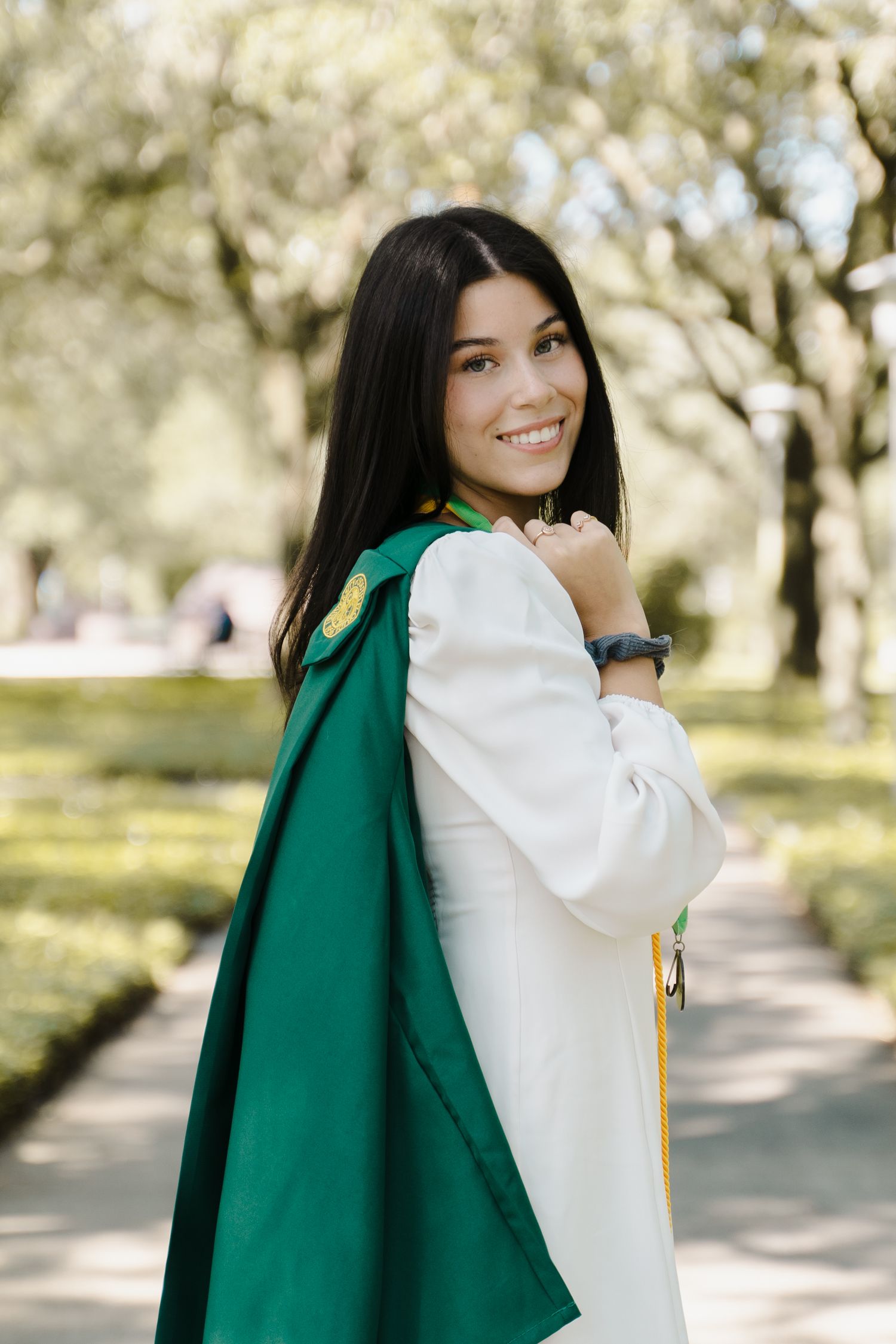 Smiling young woman in white graduation gown with green hood posing outdoors in a tree-lined campus setting.