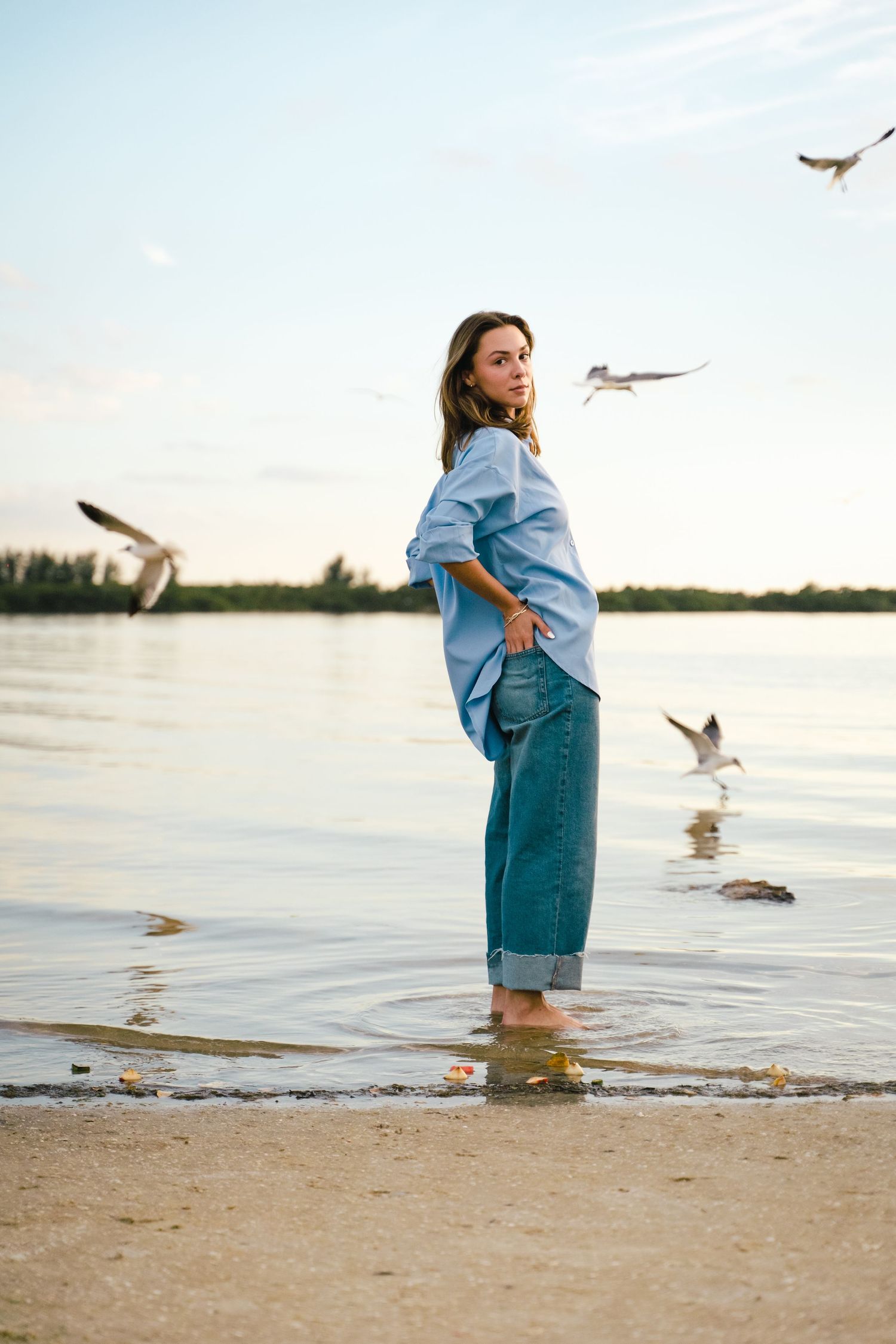 Woman in blue shirt and jeans standing barefoot in shallow water with seagulls flying around her.