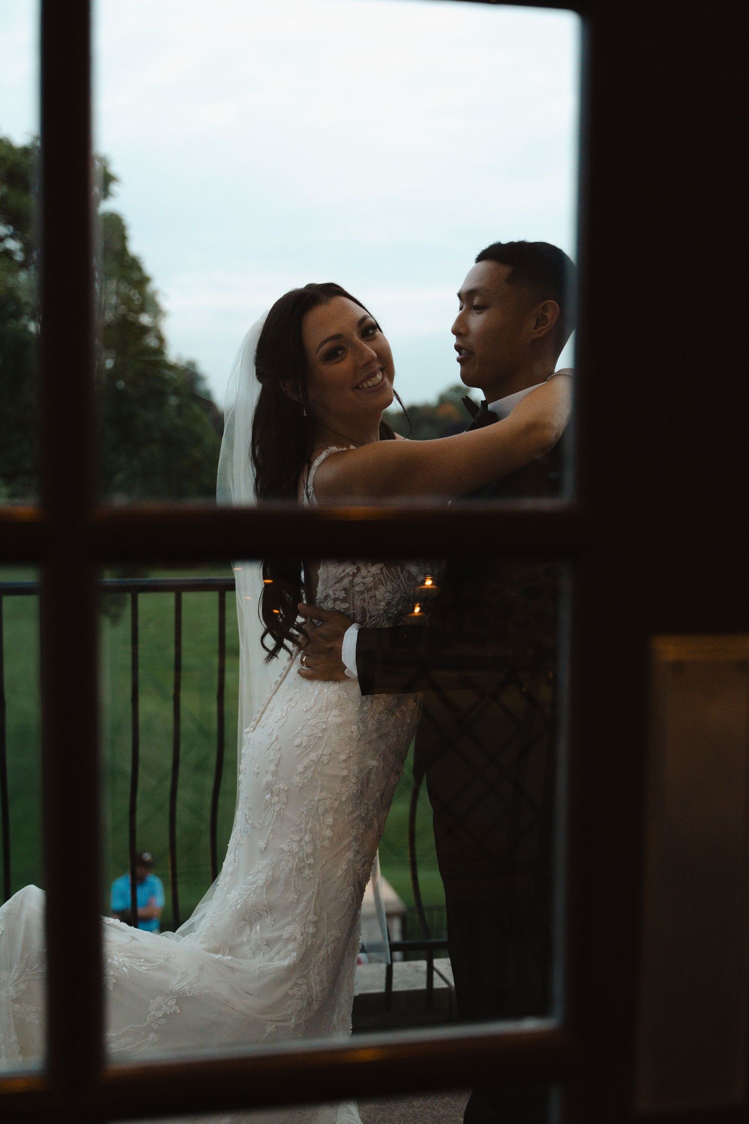 Bride and groom embrace on balcony overlooking golf course, photographed through window.