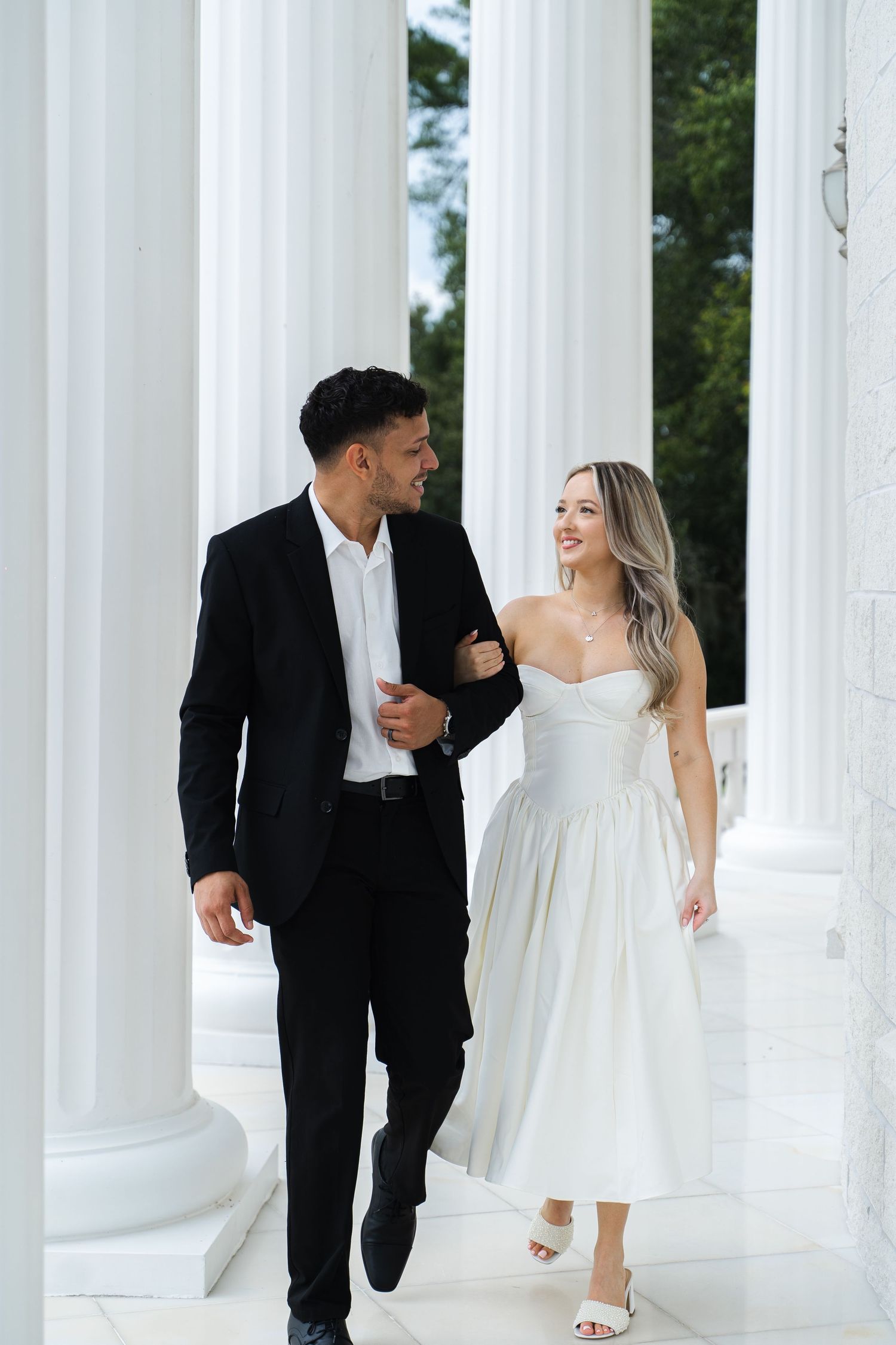 Happy couple walking arm in arm between white columns, bride in white midi dress and groom in black suit.