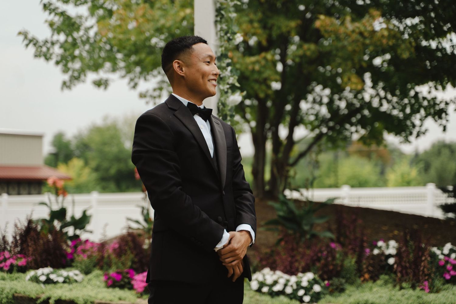 Handsome groom in black tuxedo and bow tie smiles outdoors before wedding ceremony.