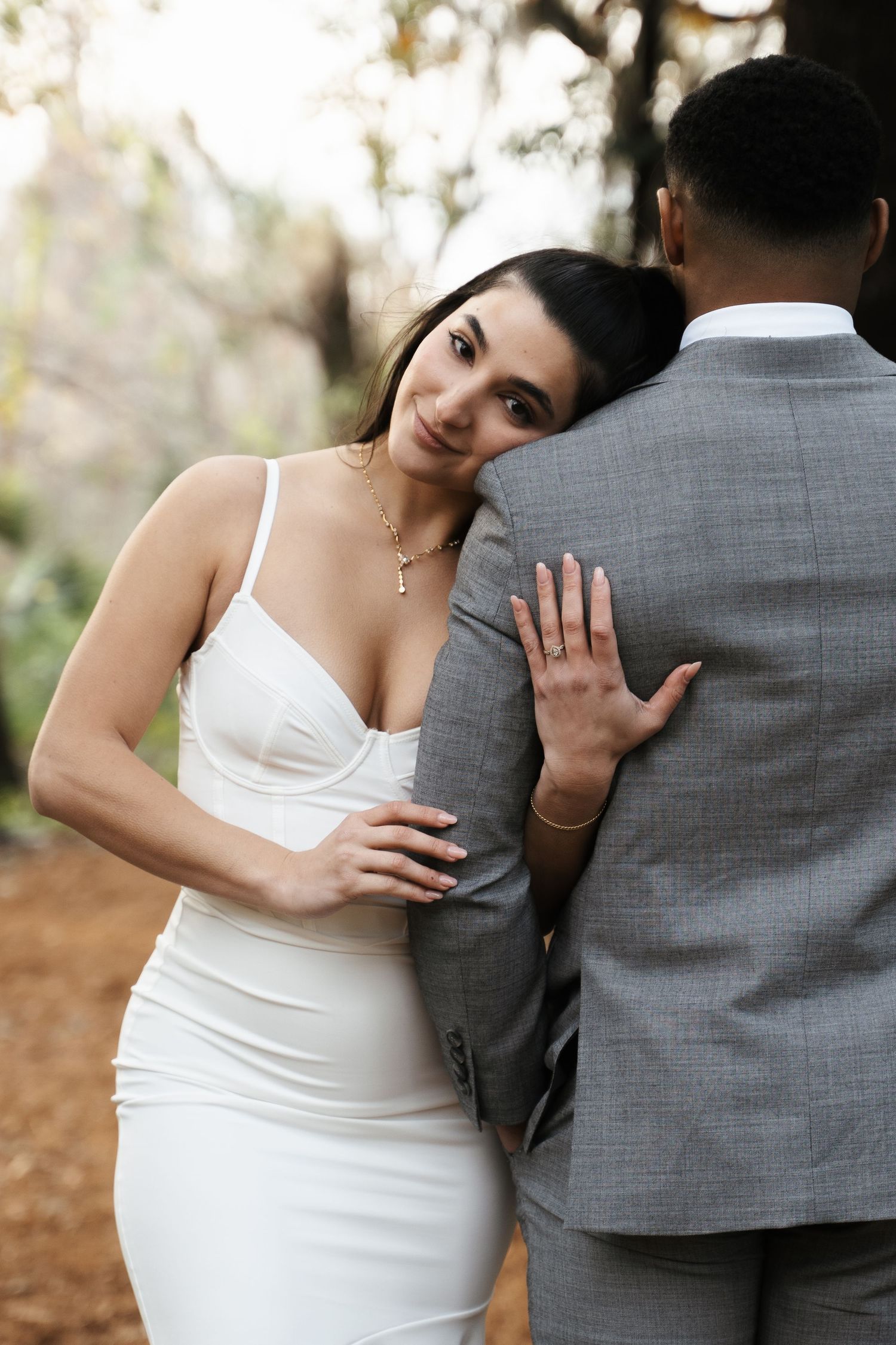Bride in fitted white dress leaning on grooms back, showing engagement ring in wooded setting.