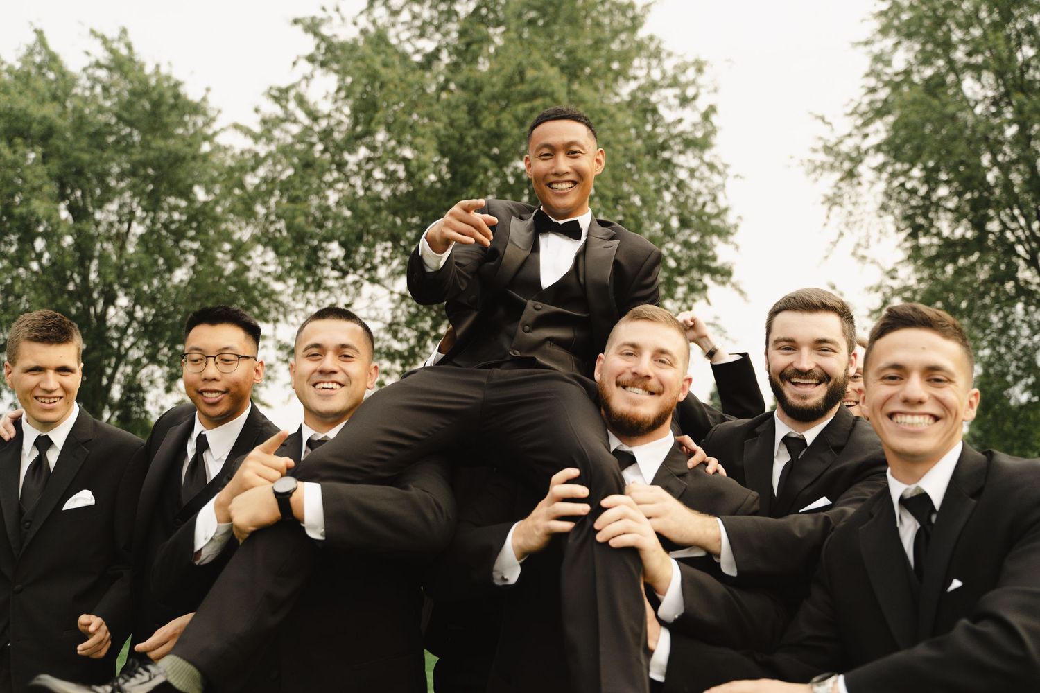 Groom being lifted by smiling groomsmen all dressed in black tuxedos outdoors.