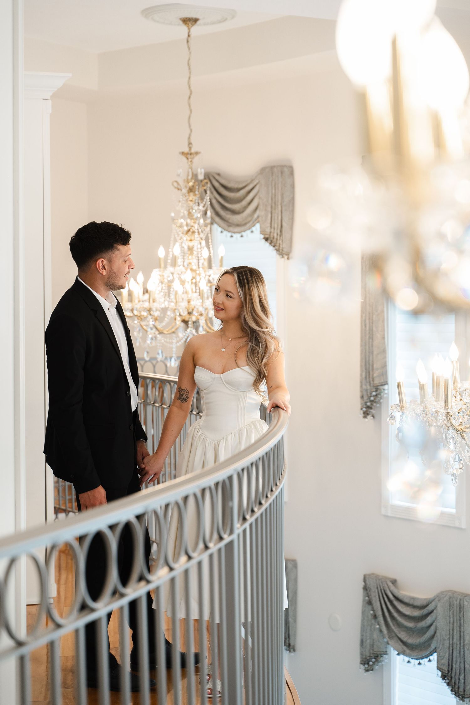 Bride and groom holding hands on a curved staircase balcony beneath an elegant chandelier.