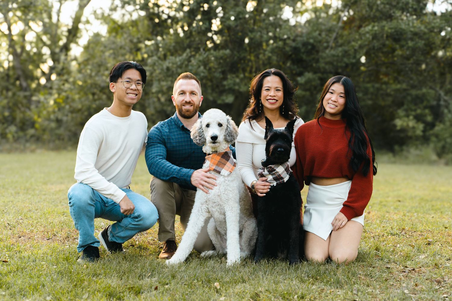 Family of four posing with two dogs on grass in a sunny outdoor park setting.