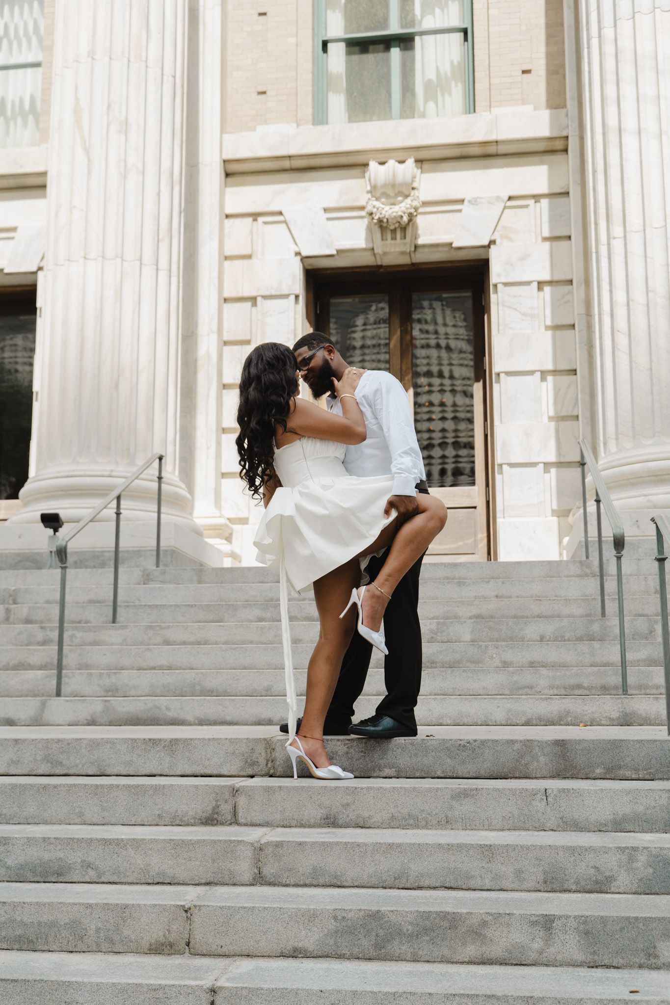 A Black couple shares a romantic kiss on marble steps of a grand building, woman lifted by partner.