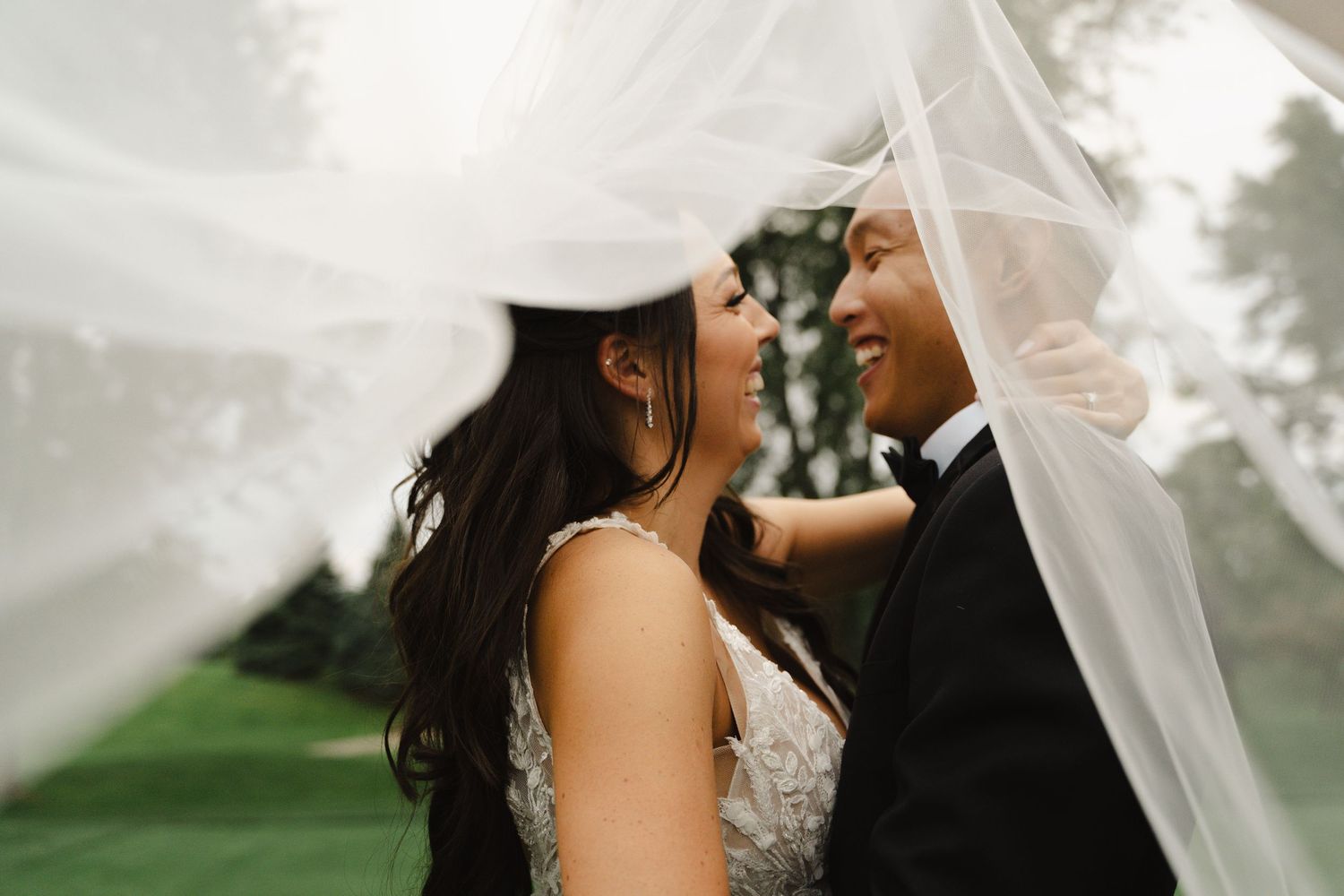 Bride and groom share a romantic moment under a flowing white veil, smiling at each other.