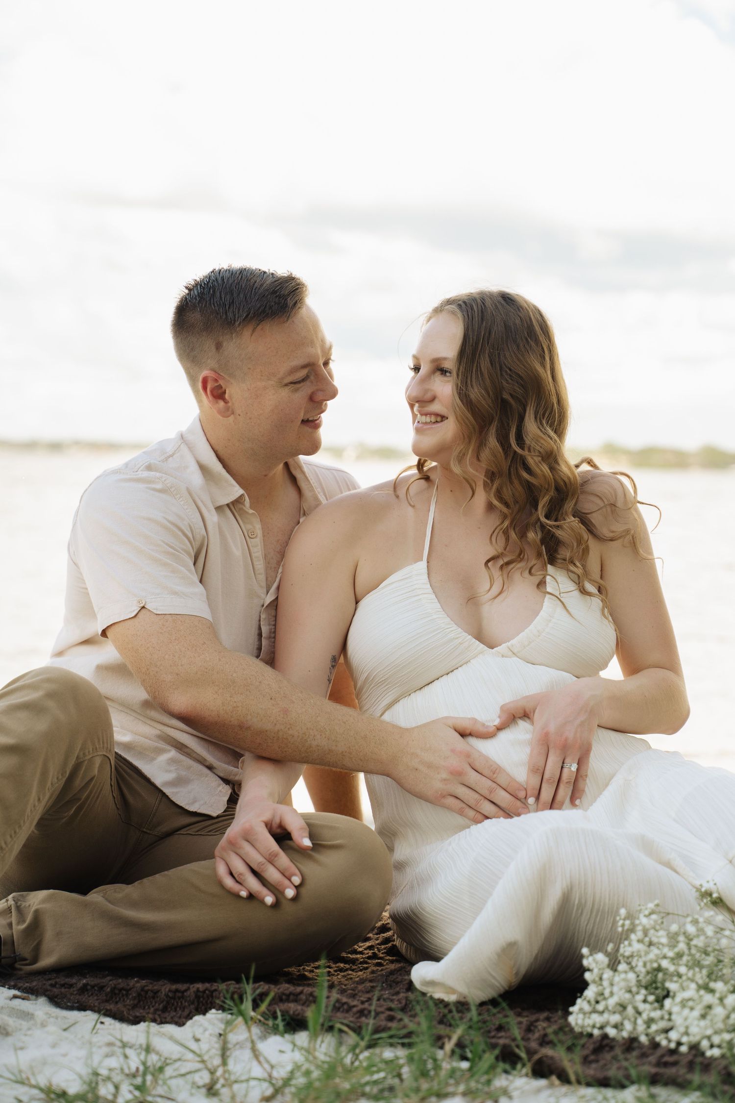 Happy couple sitting outdoors making heart shape on pregnant belly near water with baby's breath flowers nearby.
