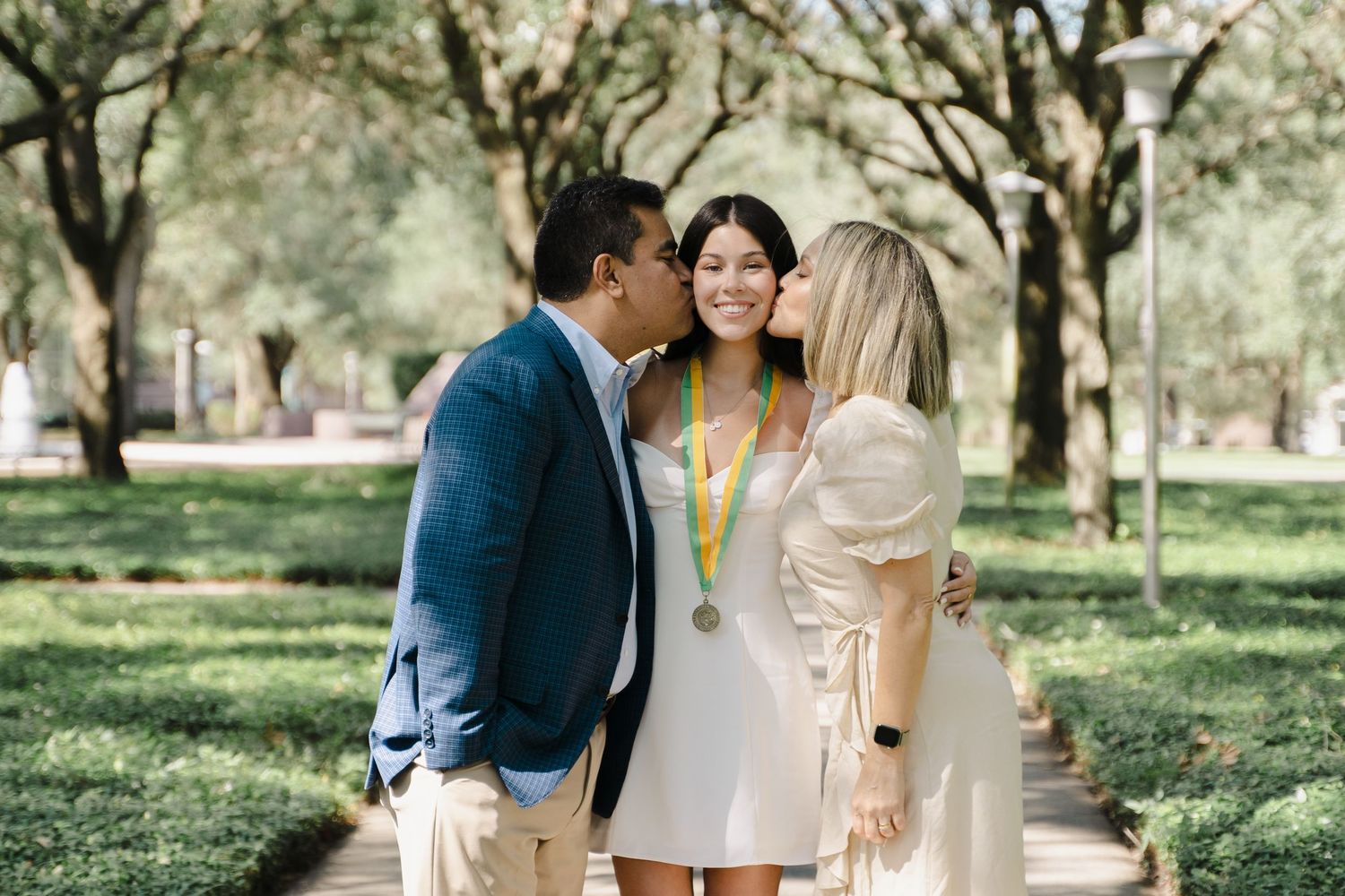 Proud graduate in white dress receives kisses from parents on tree-lined campus walkway.