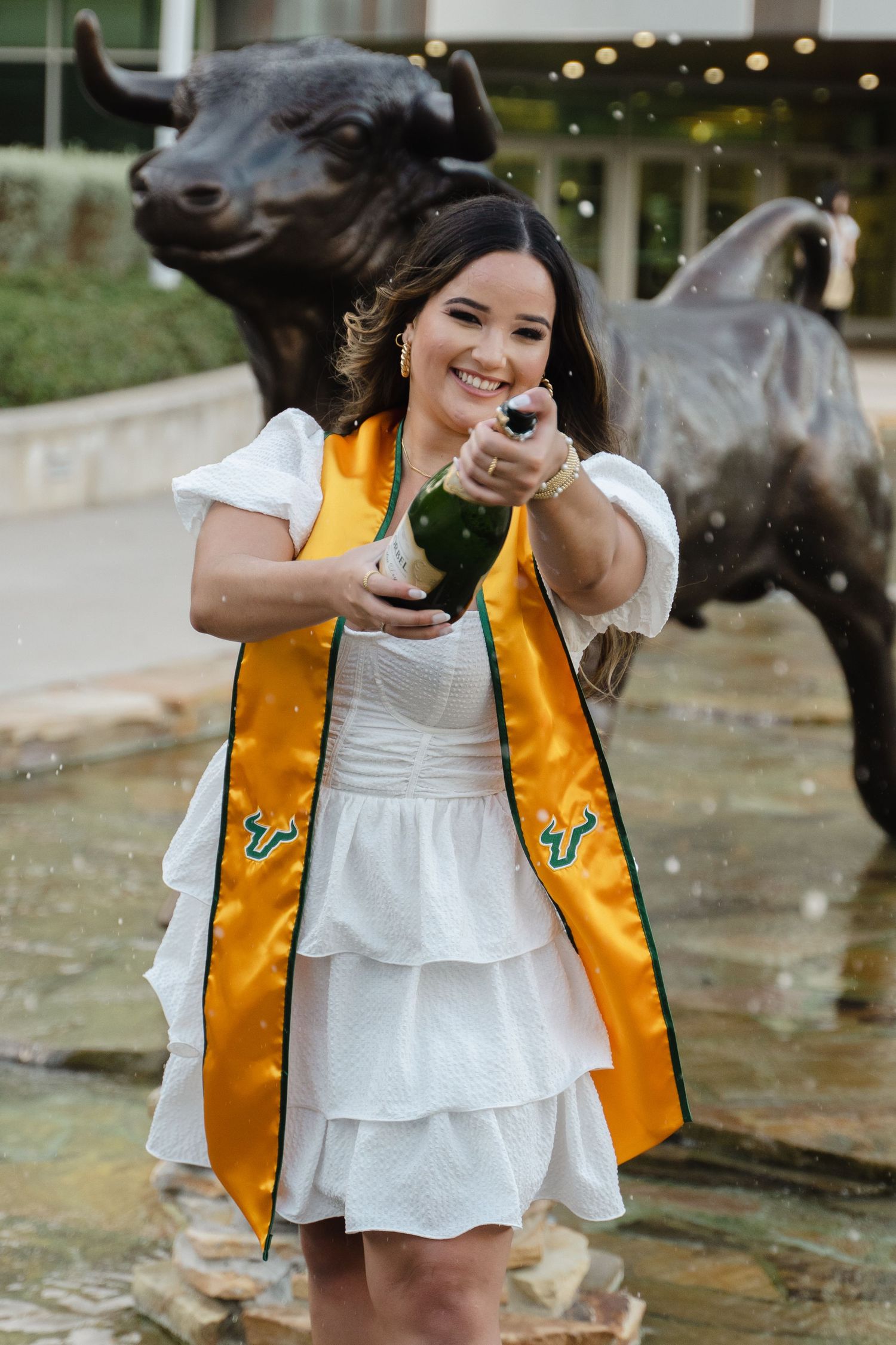 Excited graduate in white dress and gold USF stole pops champagne by bull statue.