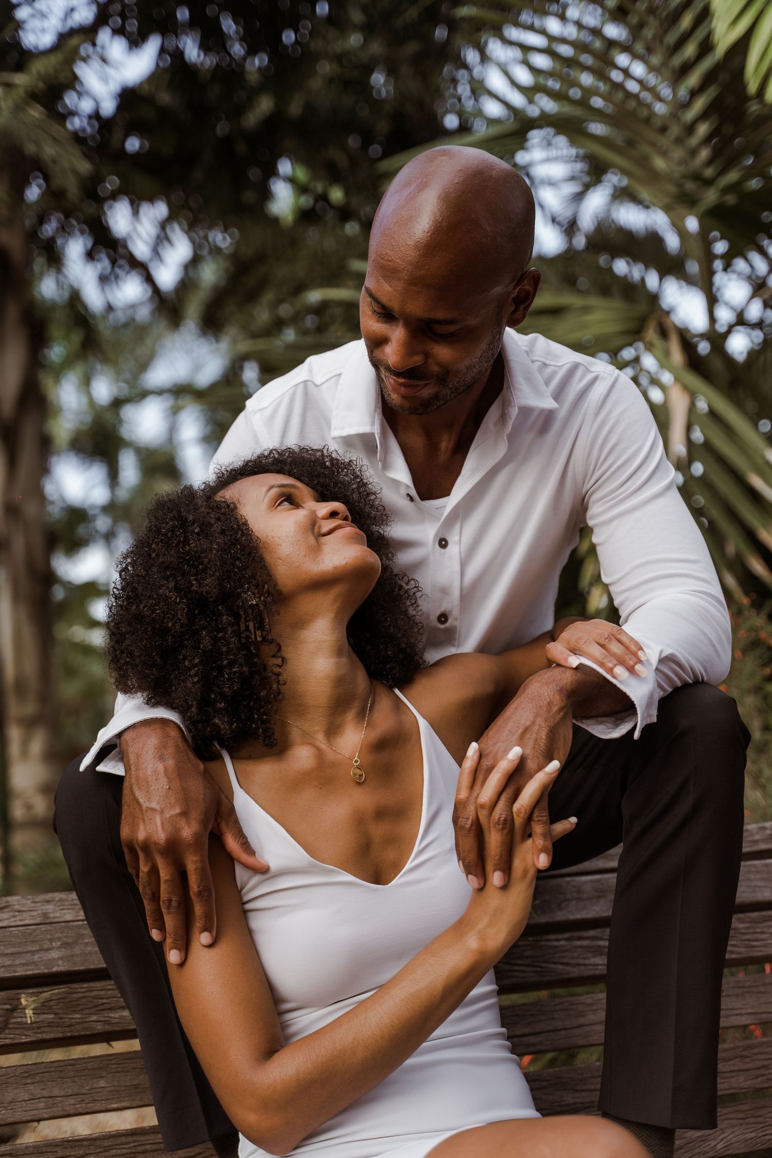 Smiling couple sitting on a wooden bench in a lush tropical park setting.