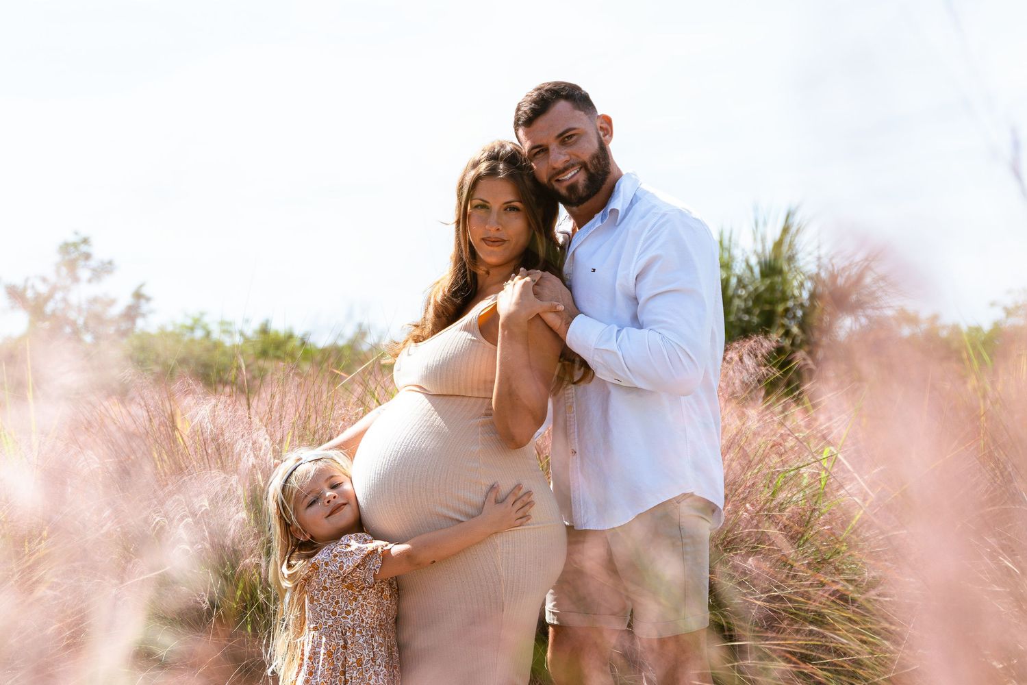 Happy family maternity photo with pregnant mother, father, and young daughter in pink grass field.