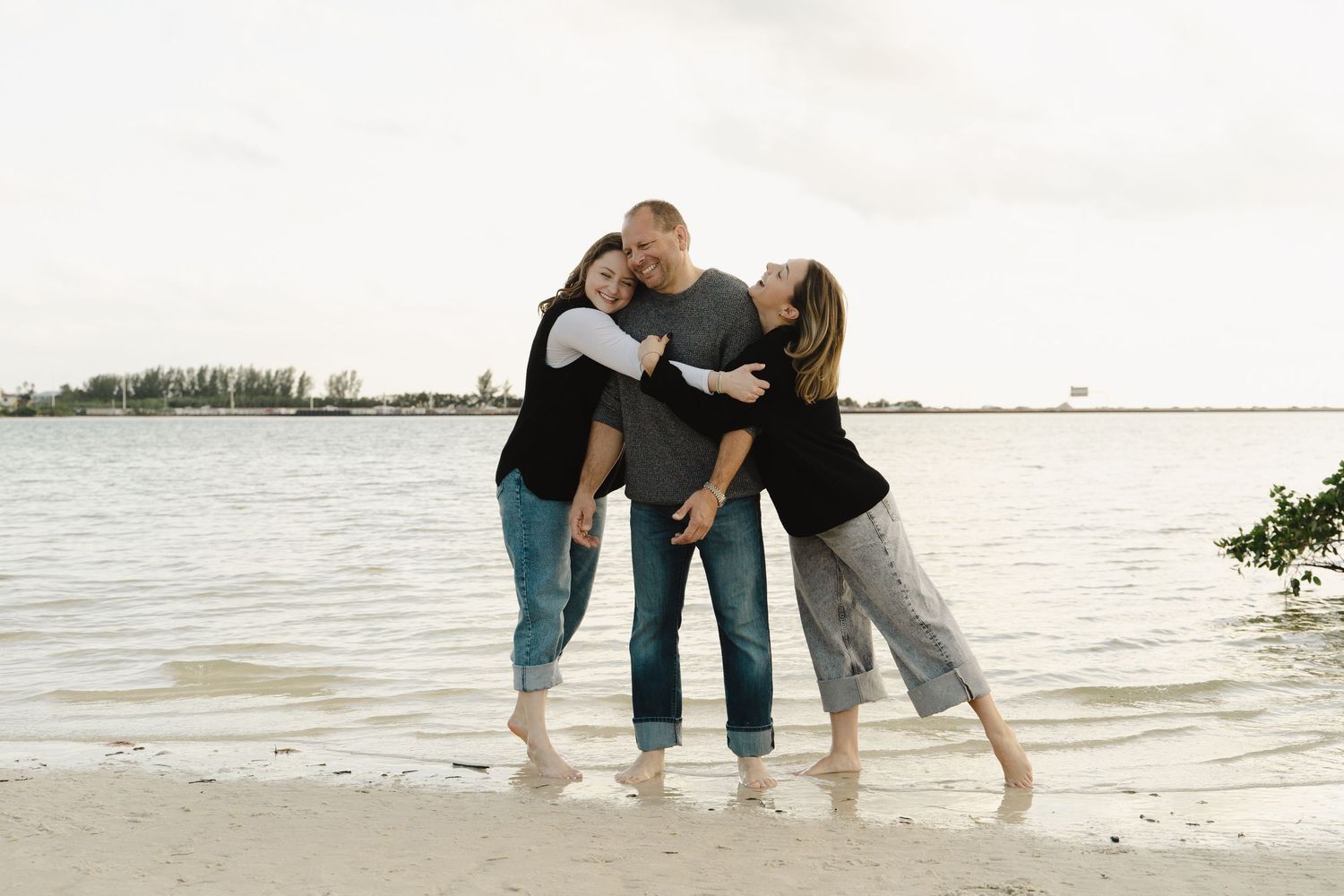 Family of three laughing and embracing on a sandy beach shoreline at sunset.