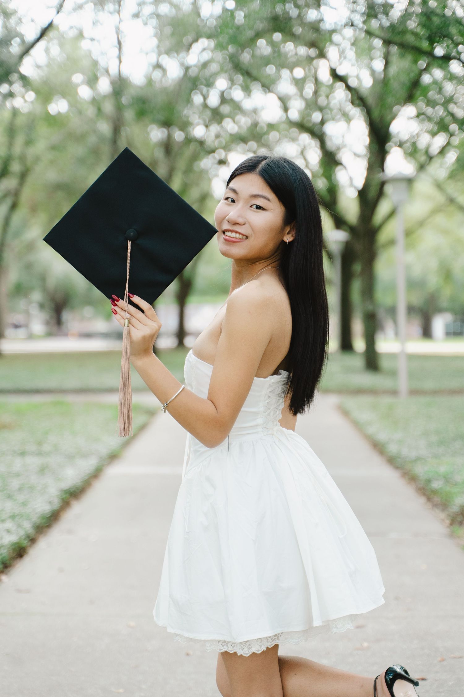 Smiling young woman in white dress holding graduation cap outdoors in tree-lined pathway.