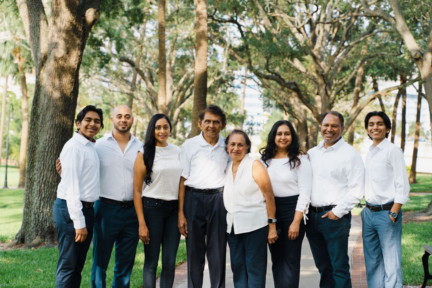 Group of eight smiling family members wearing white shirts and dark pants posing together under large oak trees.
