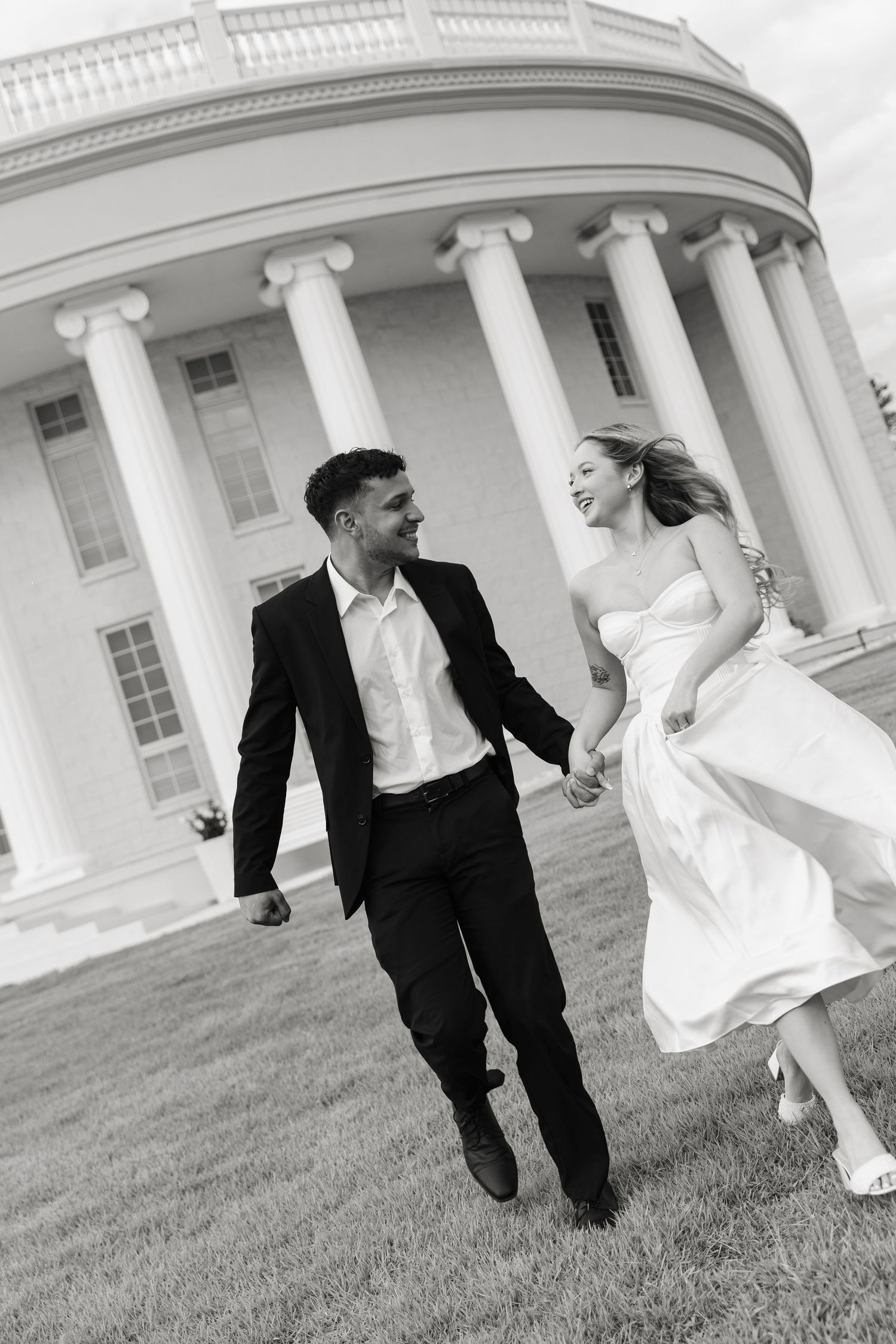 Black and white photo of laughing newlyweds running hand in hand outside columned wedding venue.