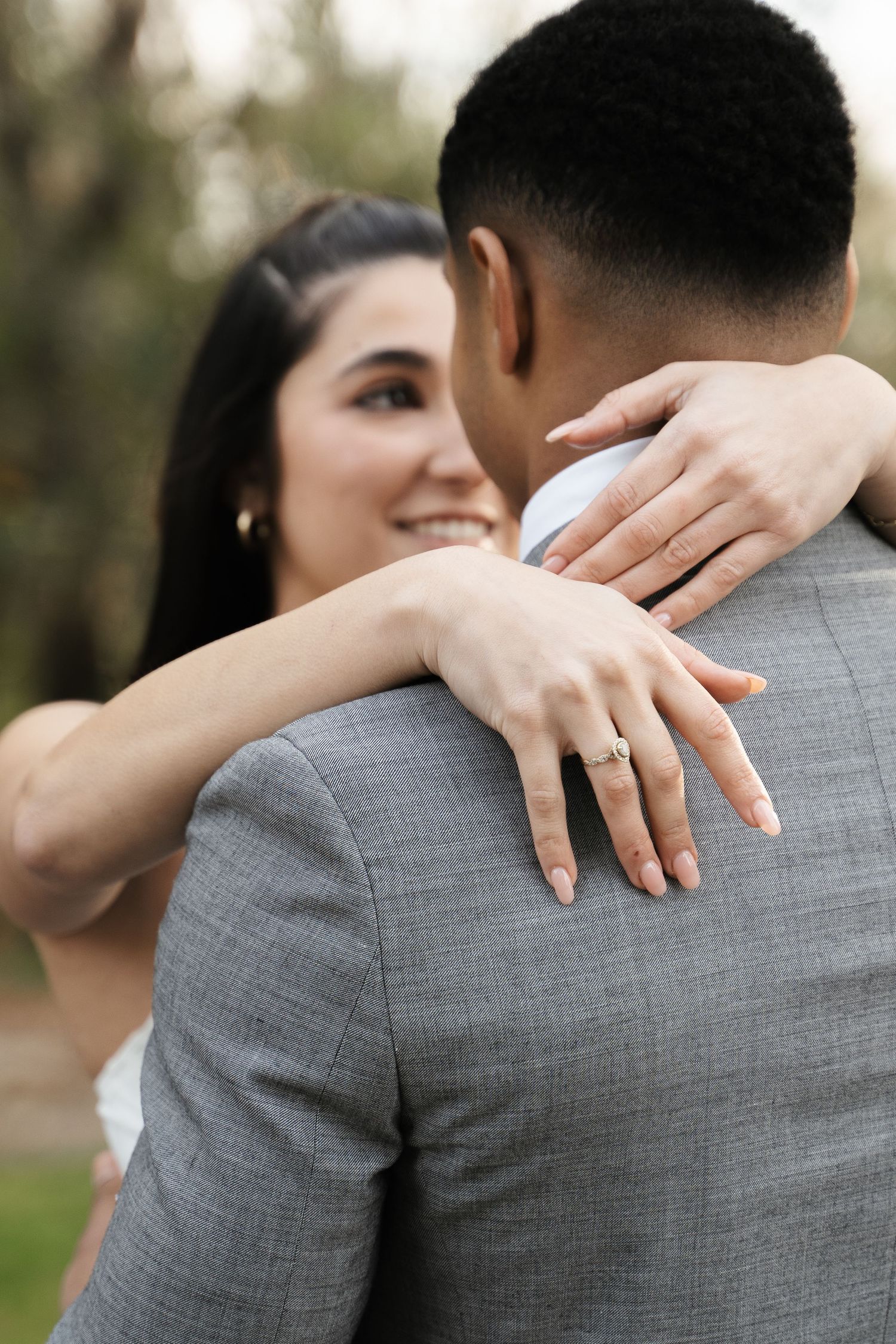 Smiling bride embracing groom from behind showing diamond engagement ring on her hand outdoors.