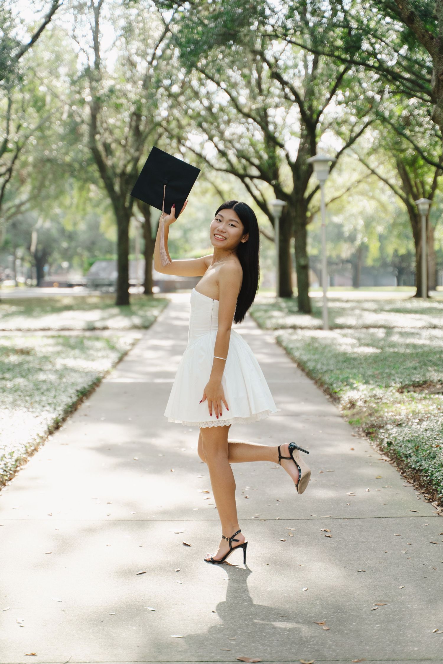 A joyful young woman in a white dress holds her graduation cap while posing playfully on a sunlit campus pathway.