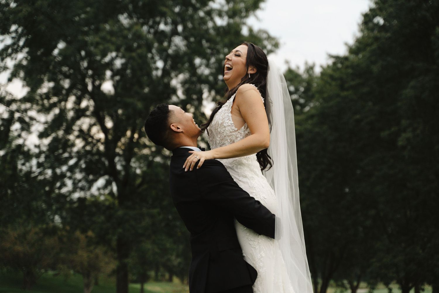 Groom lifts laughing bride in lush green park during romantic outdoor wedding portrait.