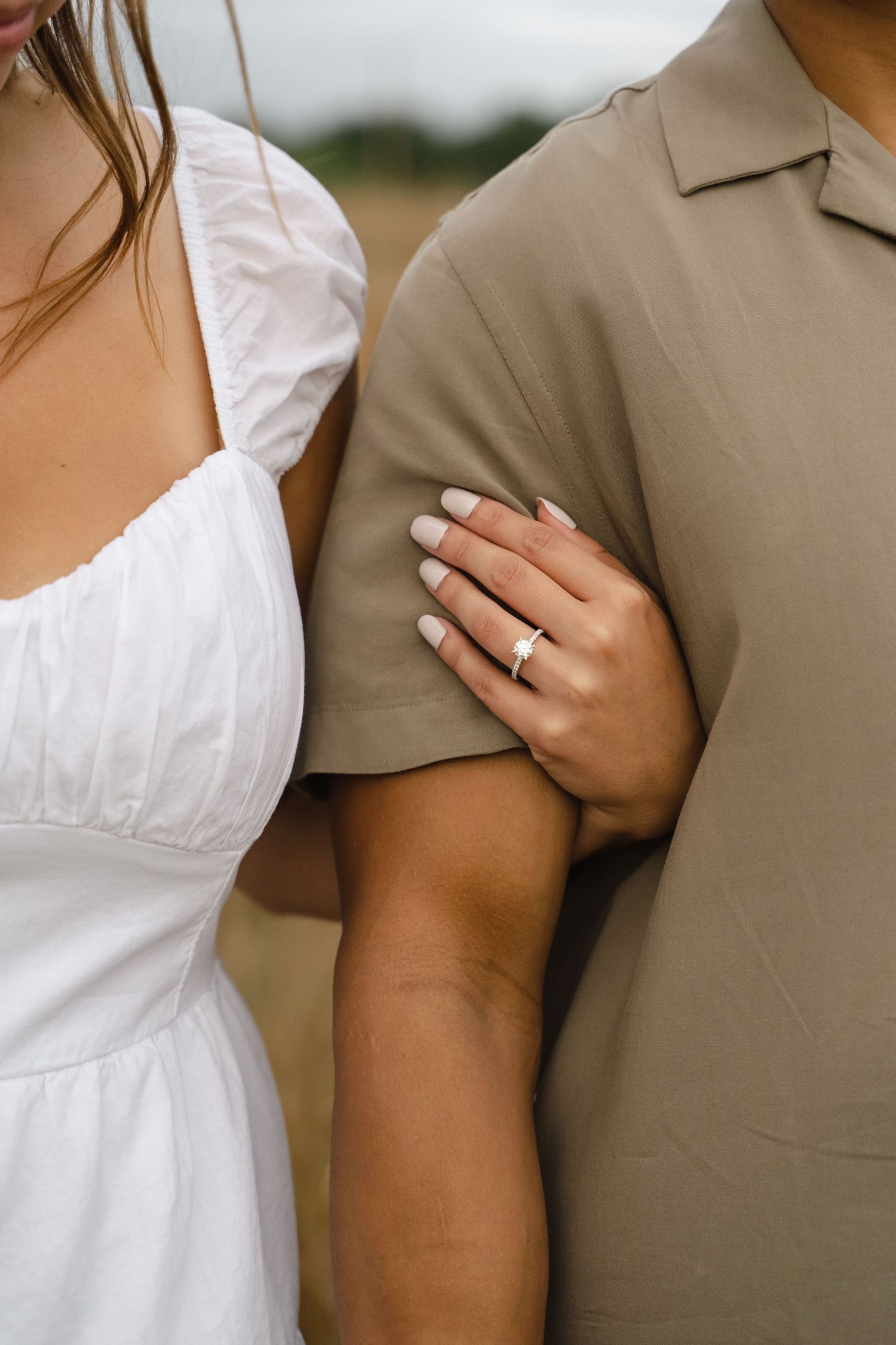Close-up of a woman in a white dress holding a man's arm, showcasing an engagement ring on her finger.