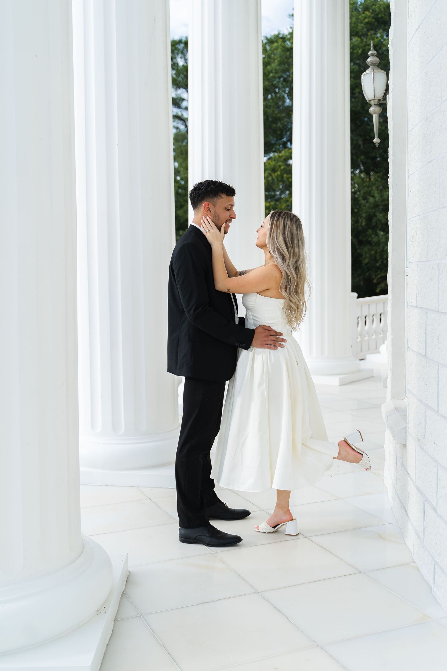 Couple sharing a romantic moment between white columns, bride in white dress and groom in black suit.