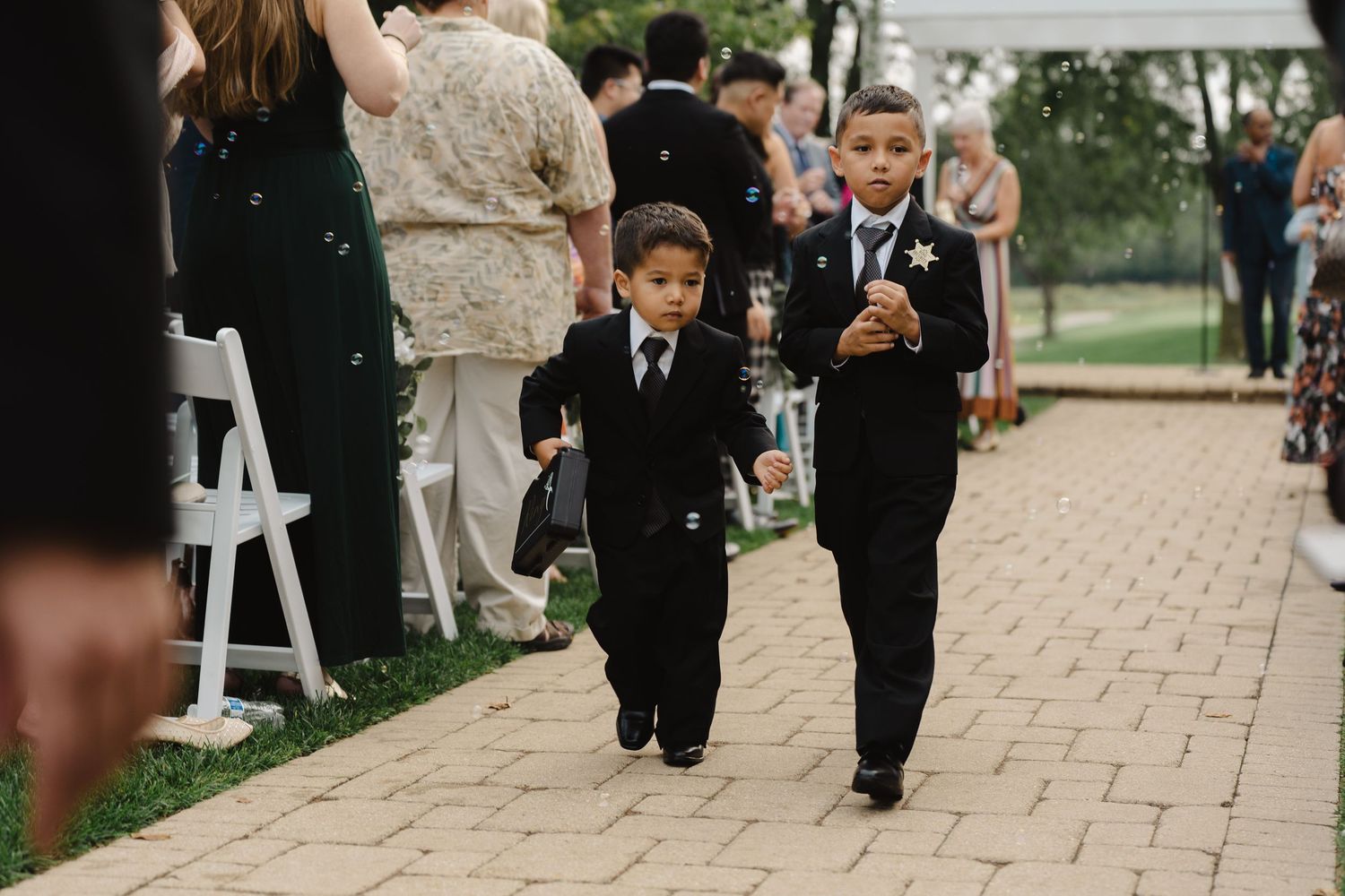 Two young ring bearers in black suits walk down brick wedding aisle blowing bubbles.