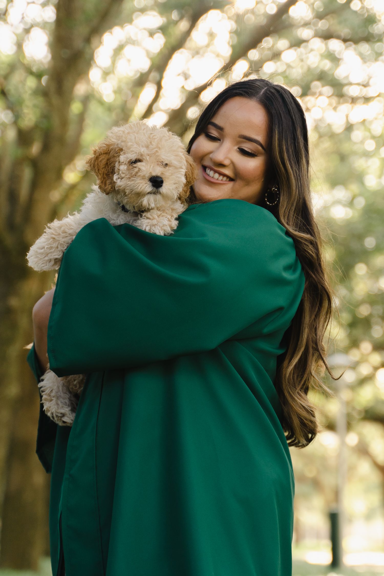 Smiling graduate in green gown holds fluffy cream puppy in sunlit park setting.