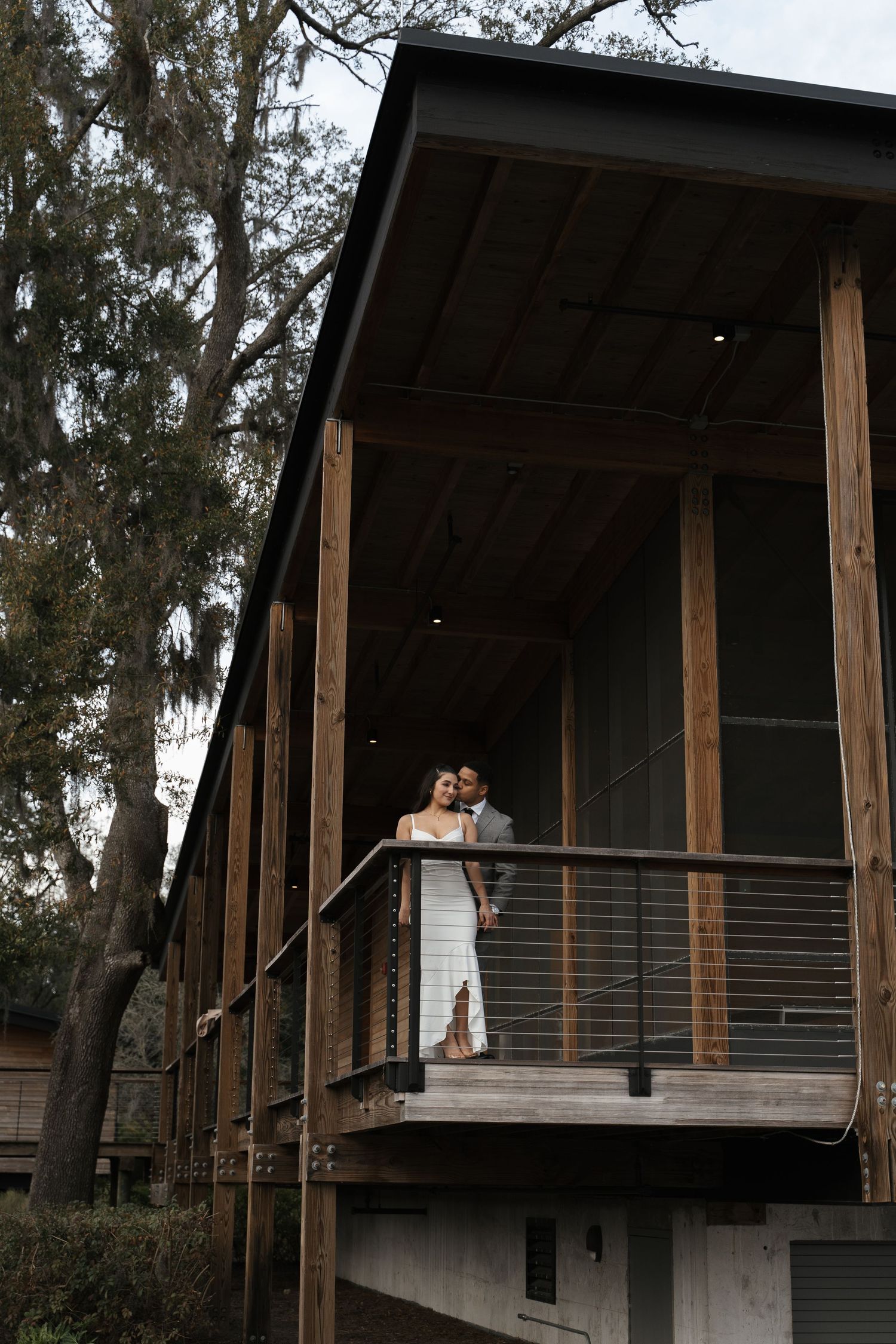 Wedding couple shares a tender moment on a wooden balcony of a rustic building surrounded by tall trees.