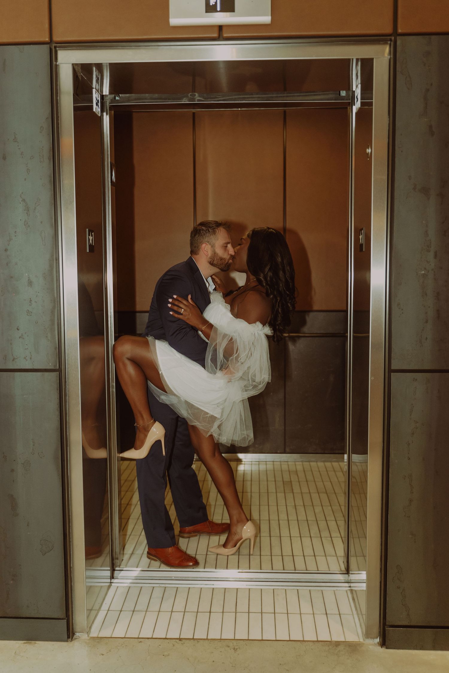 Romantic couple sharing a kiss in an elevator, groom lifting bride in white dress and heels.