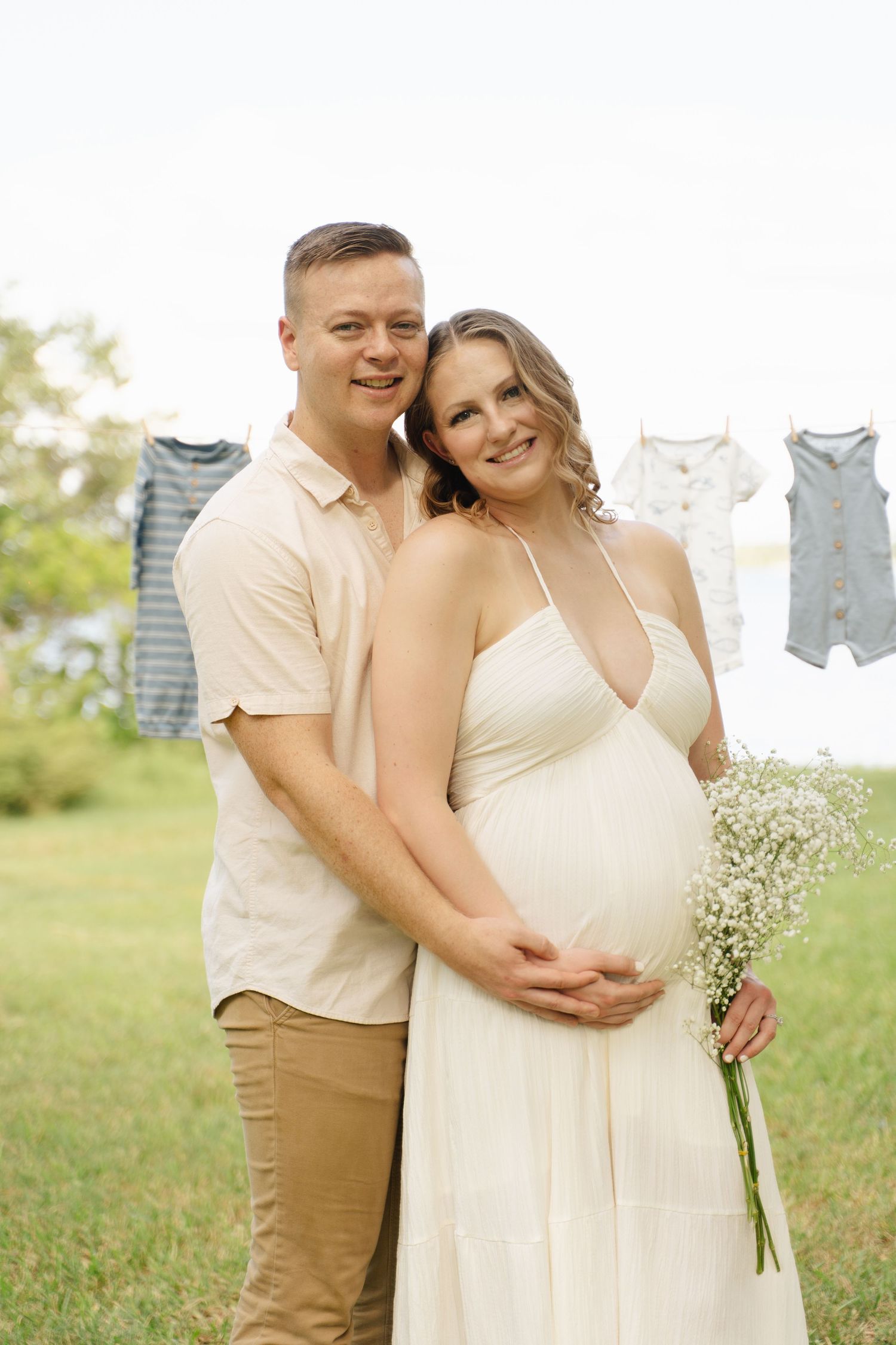 Smiling pregnant woman in white dress holding baby's breath bouquet embraced by partner outdoors with baby clothes on line.