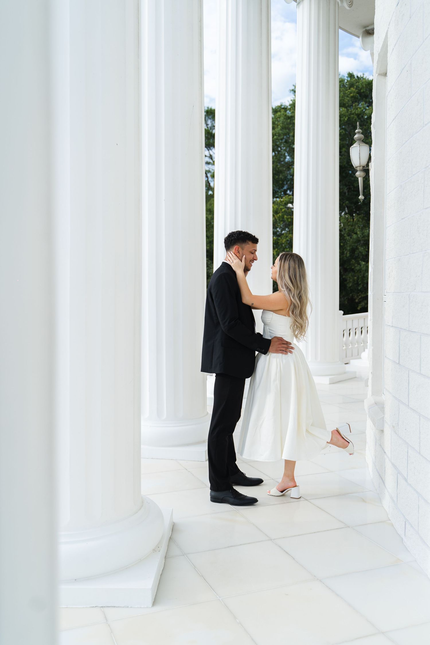 Romantic couple in wedding attire sharing an intimate moment between white neoclassical columns on a sunny day.