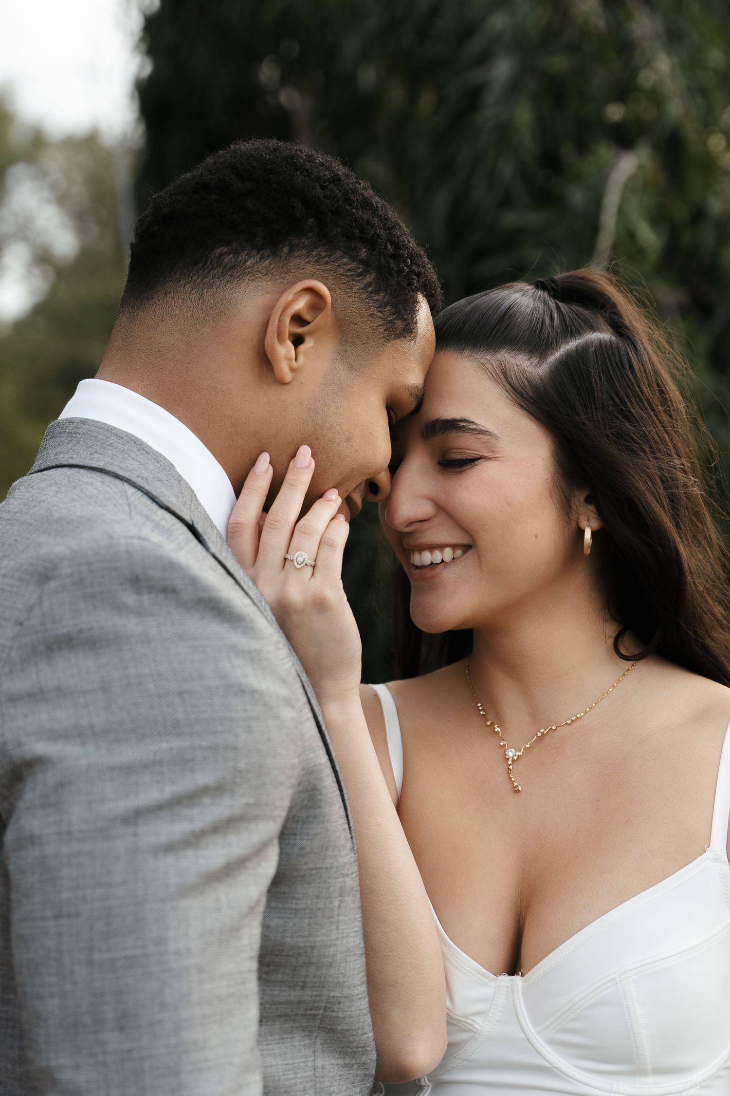 Smiling couple touching foreheads tenderly, woman in white dress with gold necklace and ring.