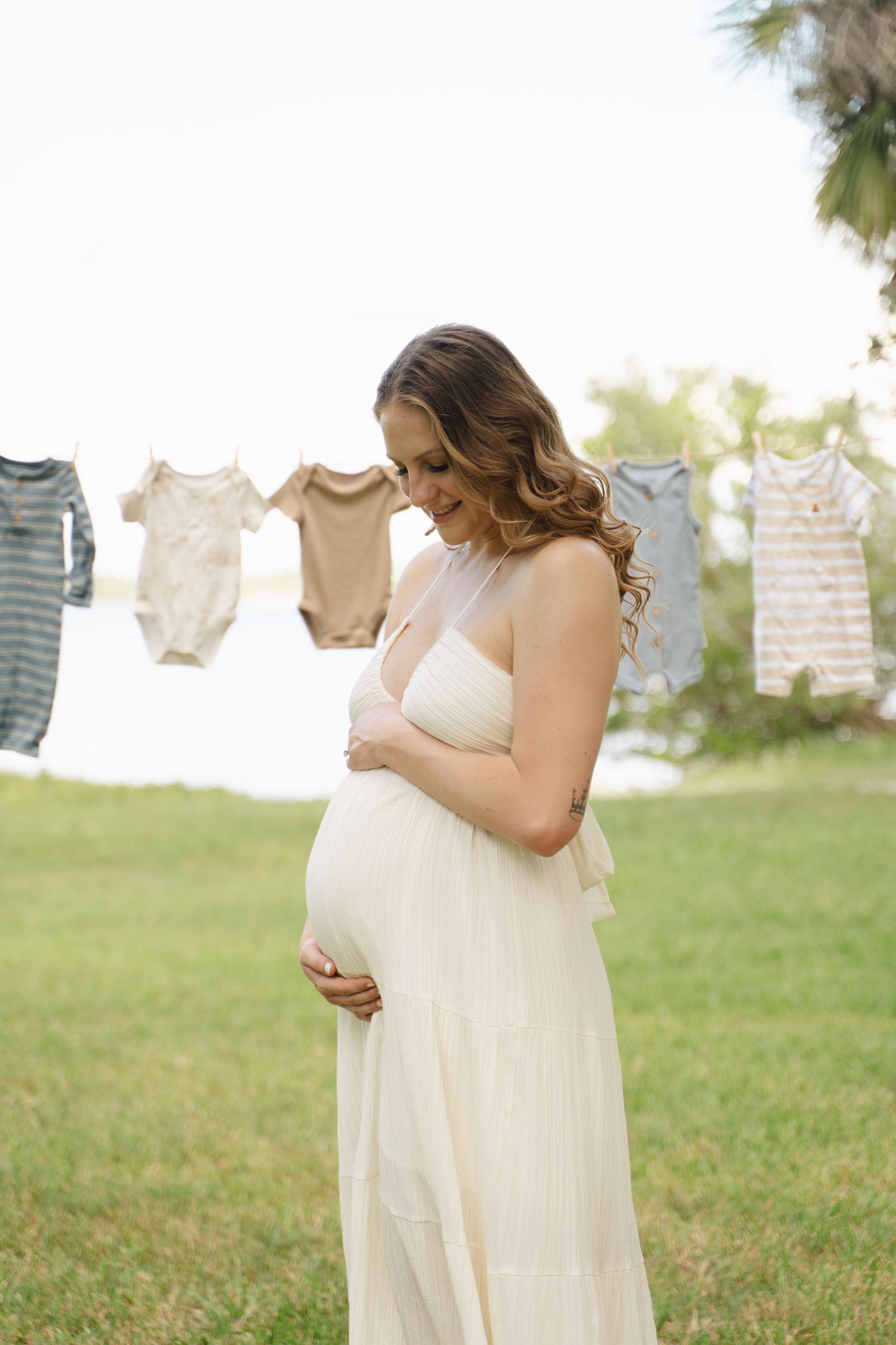 Pregnant woman in white halter dress cradling baby bump outdoors with neutral-toned baby clothes hanging on clothesline.
