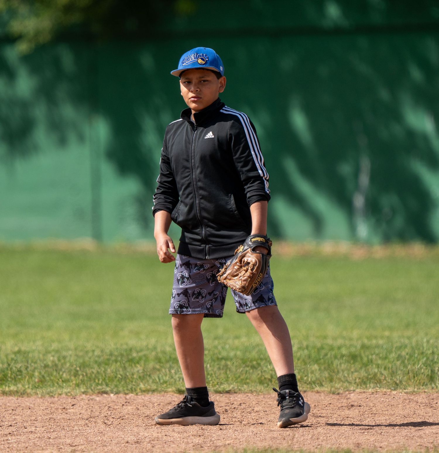 Young boy in Adidas jacket and blue cap plays baseball on dirt infield.