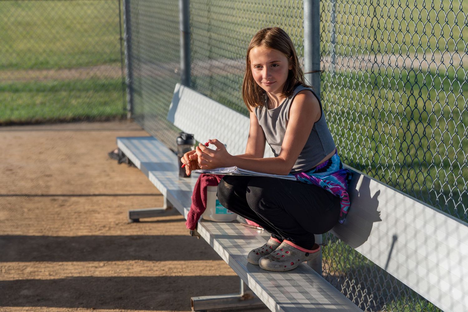 Young girl sitting on metal bleachers at a baseball field, holding a glove on a sunny day.