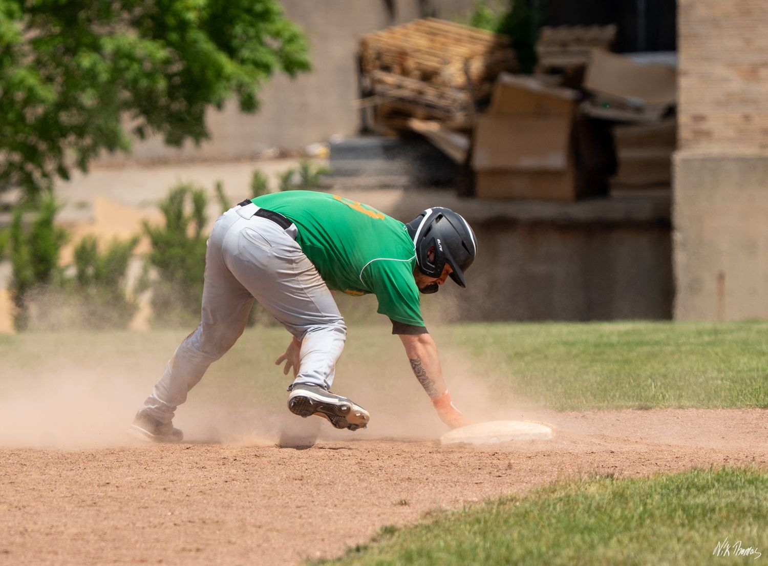 Baseball player in green jersey slides headfirst into base creating dust cloud on sunny day.