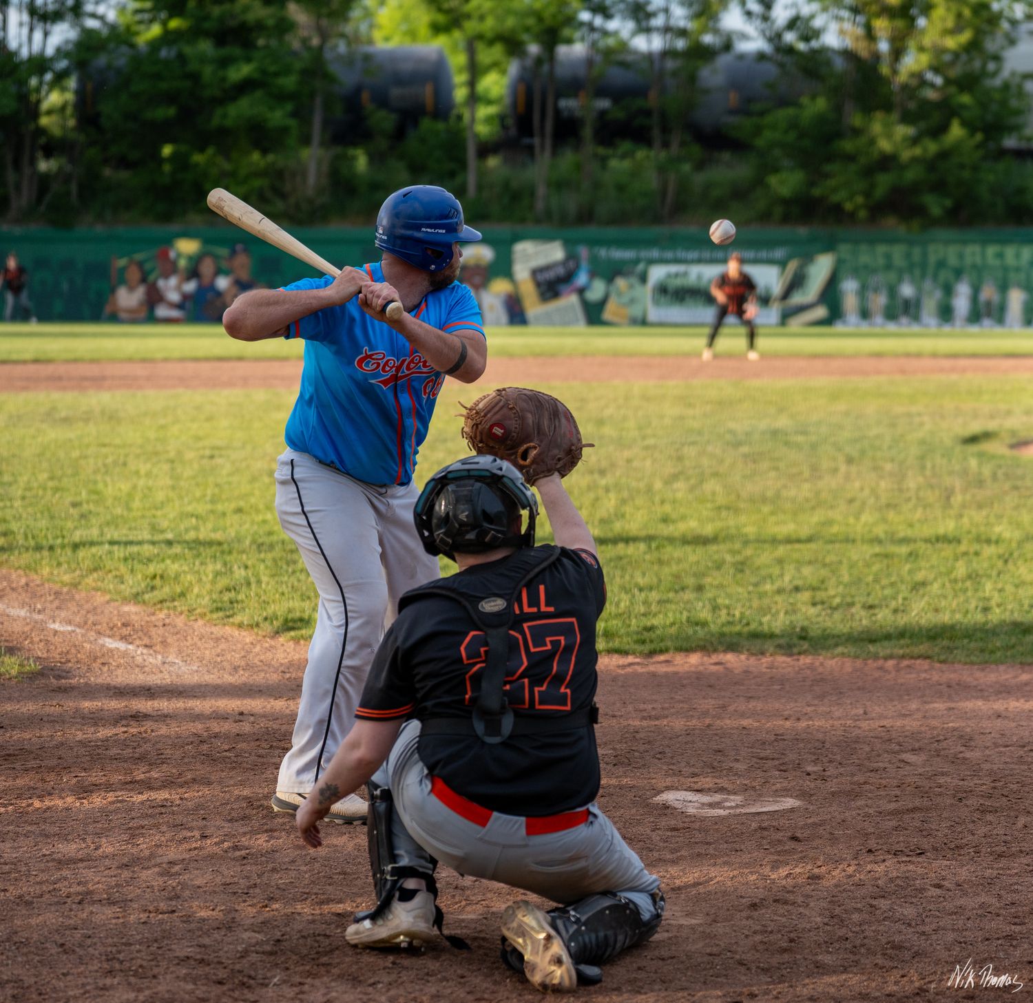 Baseball batter swings at pitch while catcher crouches behind home plate during a sunny evening game.